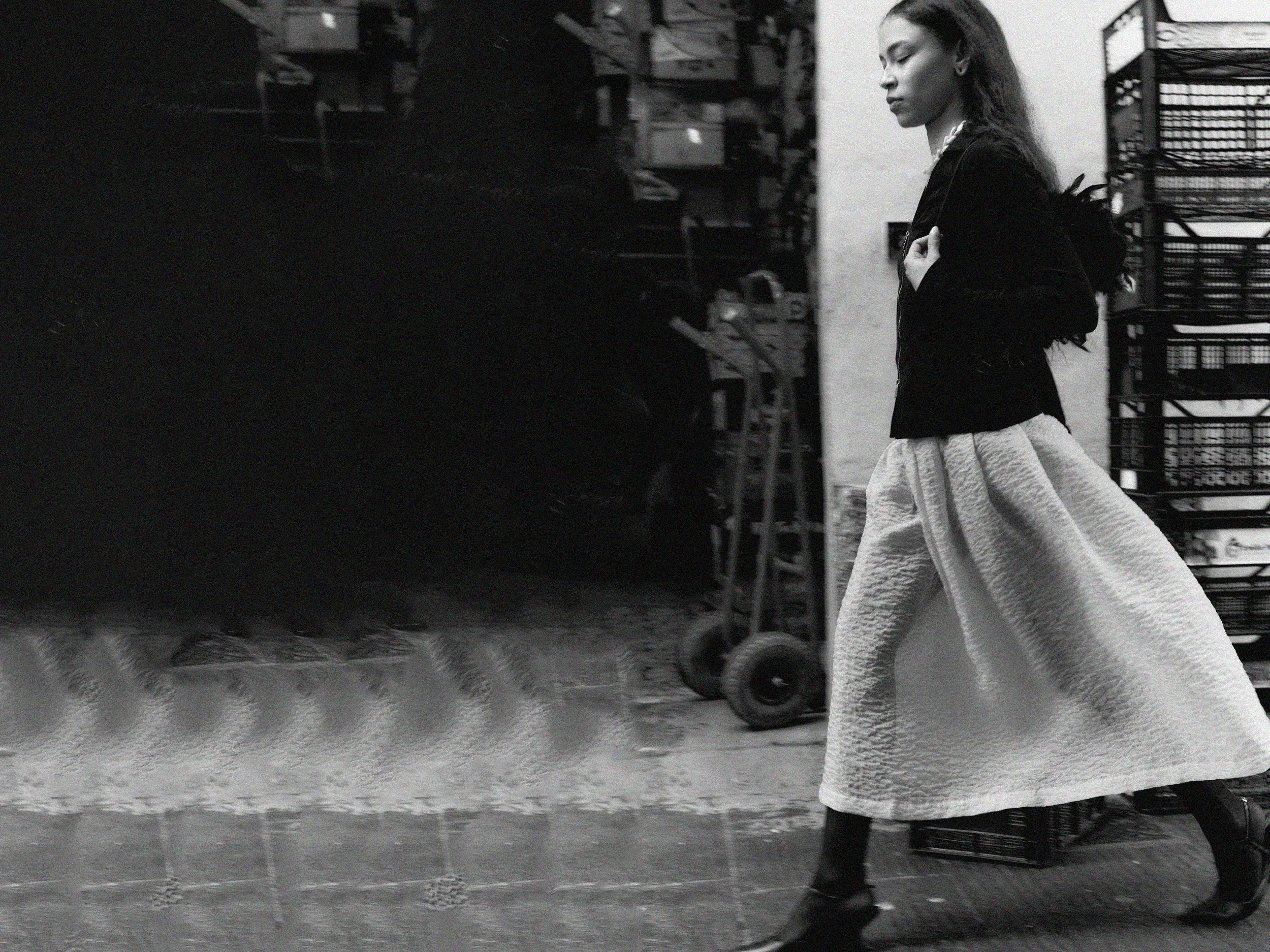 A woman wearing a long, textured skirt and a dark jacket walking past a storage or display area with metal shelves and a hand truck.
