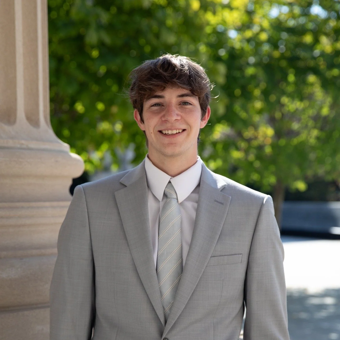 A young man smiling outdoors wearing a light gray suit and no tie. There are blurred green trees in the background.