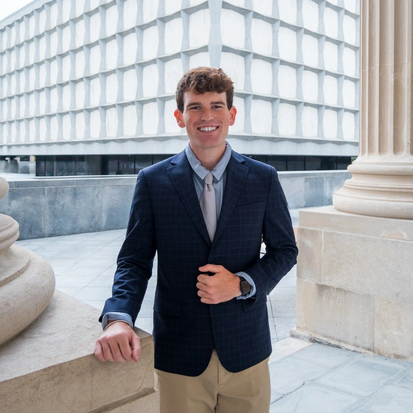 A young man with brown hair, smiling, wearing a black suit, pink shirt, and a light blue plaid tie, standing outdoors in front of a modern architectural background with white concrete patterns.
