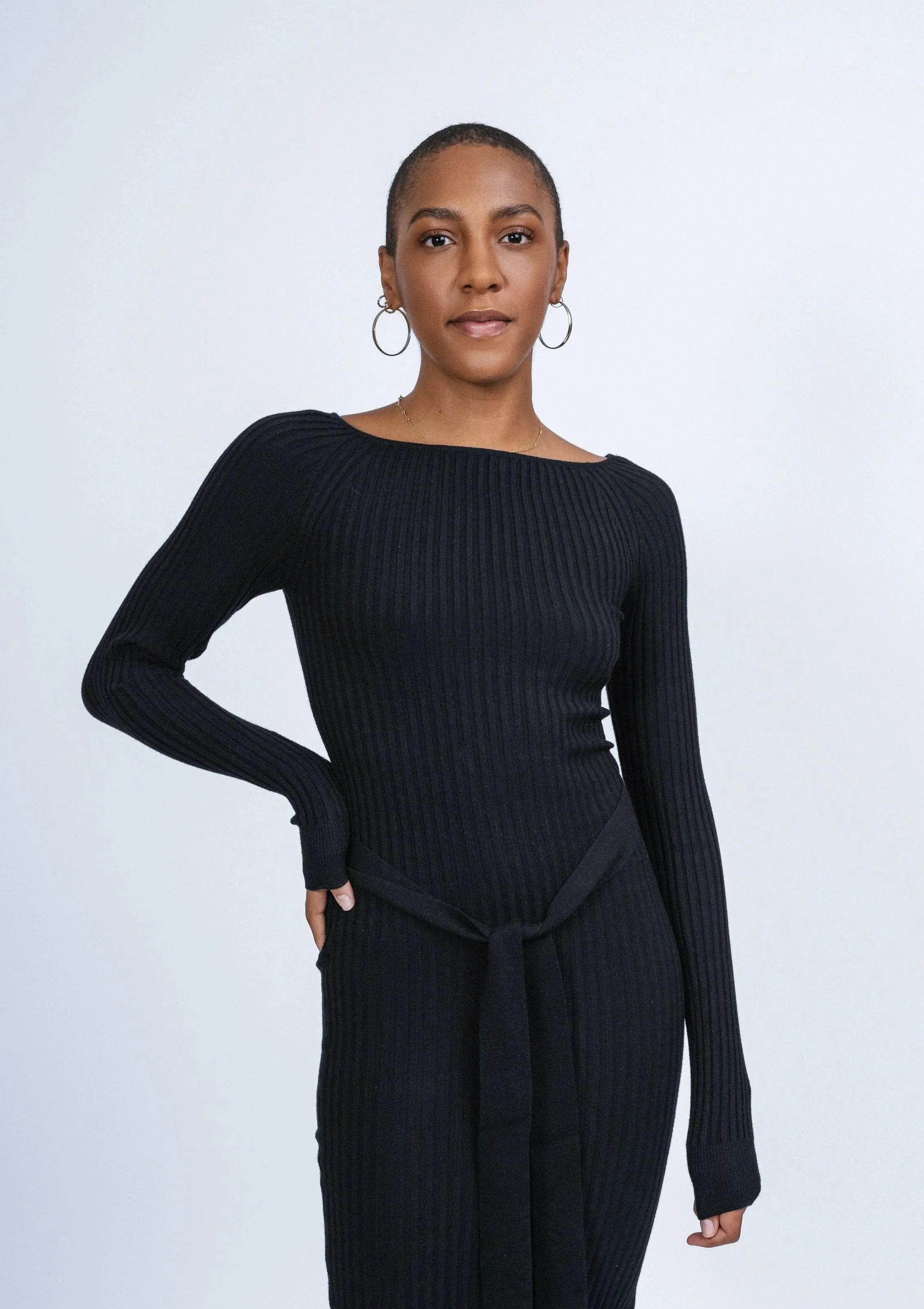 Mélanie OMAIA : Une femme debout contre un mur blanc, portant un pull noir à manches longues en maille côtelée et un pantalon noir avec une ceinture à nœud. Elle porte des boucles d'oreilles en anneaux et un collier fin.