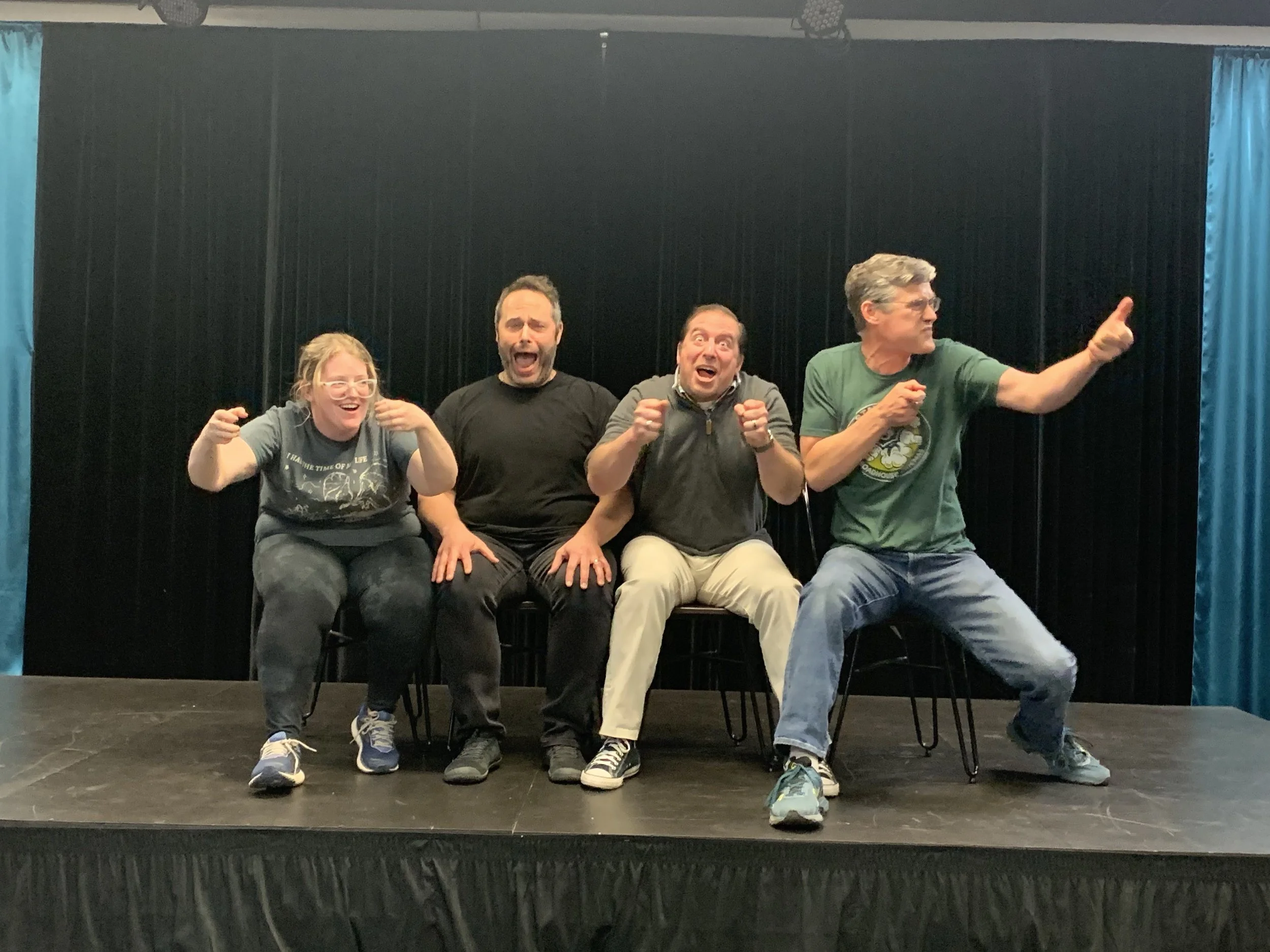 Four improv students perform an expressive scene on stage at Peak Improv Theater in Colorado Springs, using exaggerated gestures and character work during a class exercise.