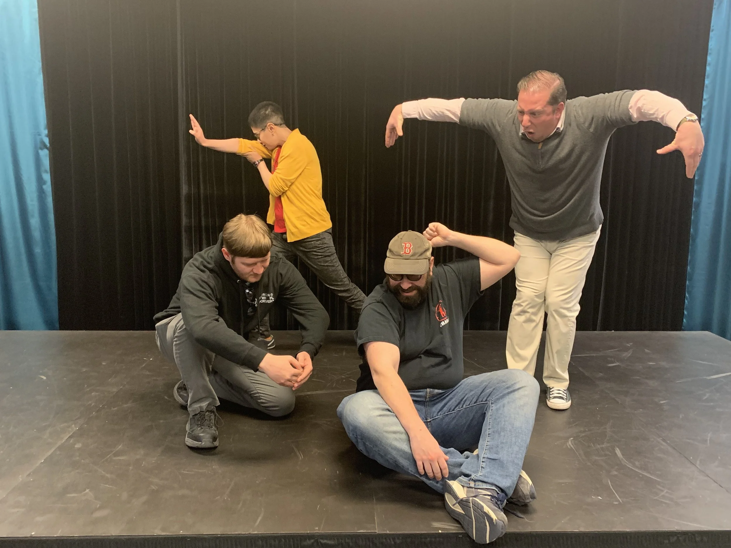 Four improv students perform a collaborative scene on stage at Peak Improv Theater in Colorado Springs, using expressive body language and character poses during a class exercise.