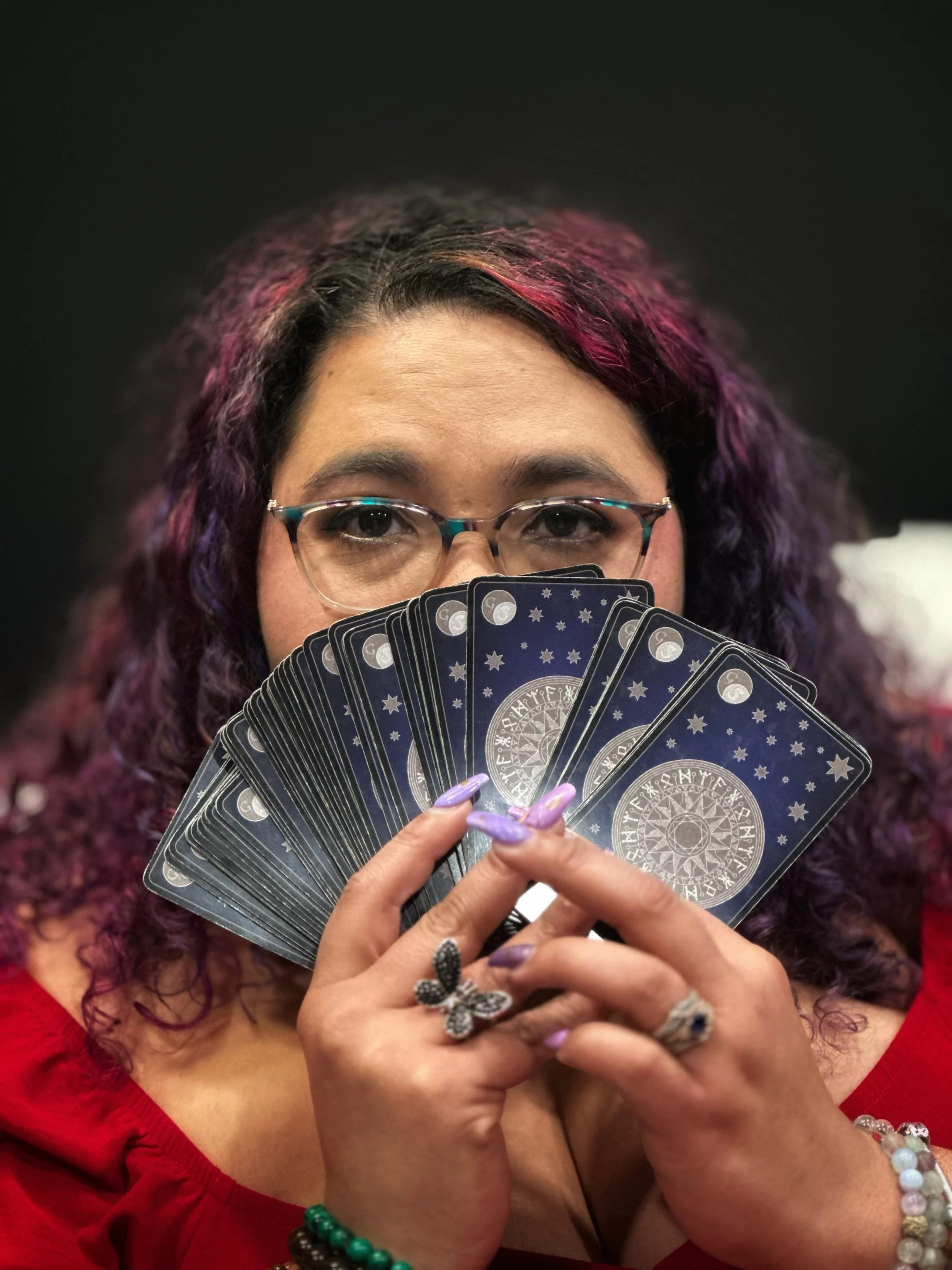 A woman with purple and pink curly hair, glasses, and wearing a red top, holding tarot cards in front of her face.