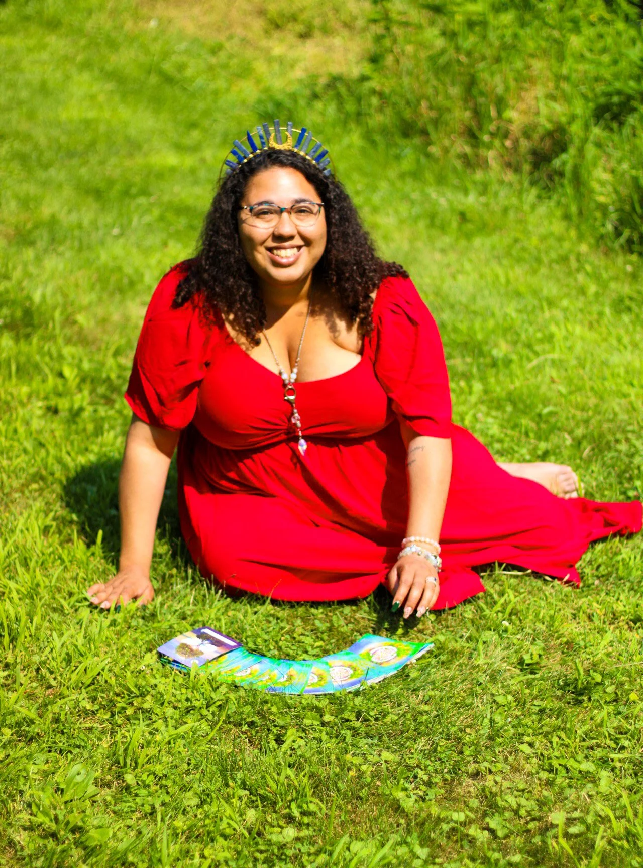 Woman in a red dress sitting on grass outdoors with tarot cards in front of her, wearing a crown and smiling.