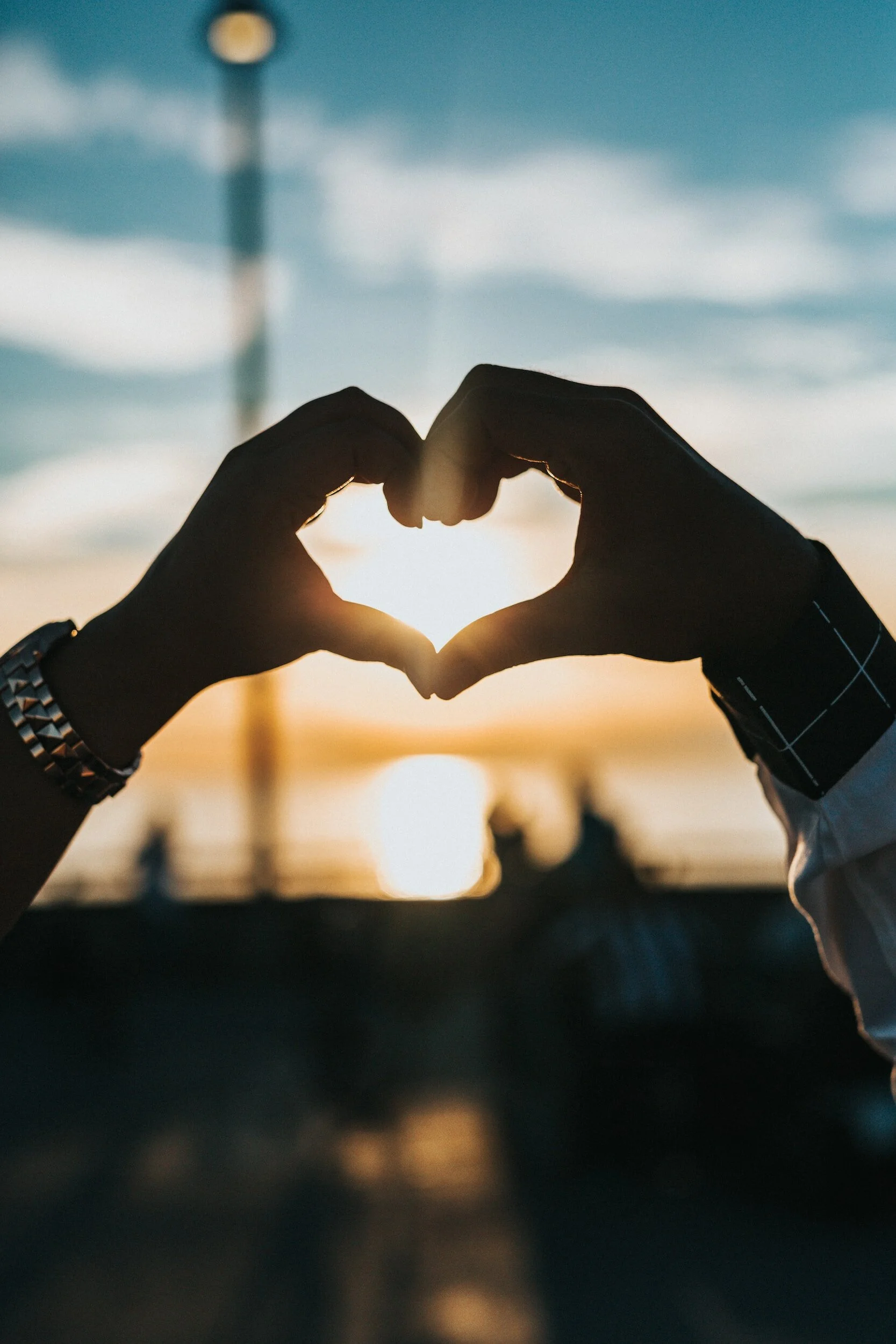 Hands forming a heart shape around the setting sun during sunset at the beach.