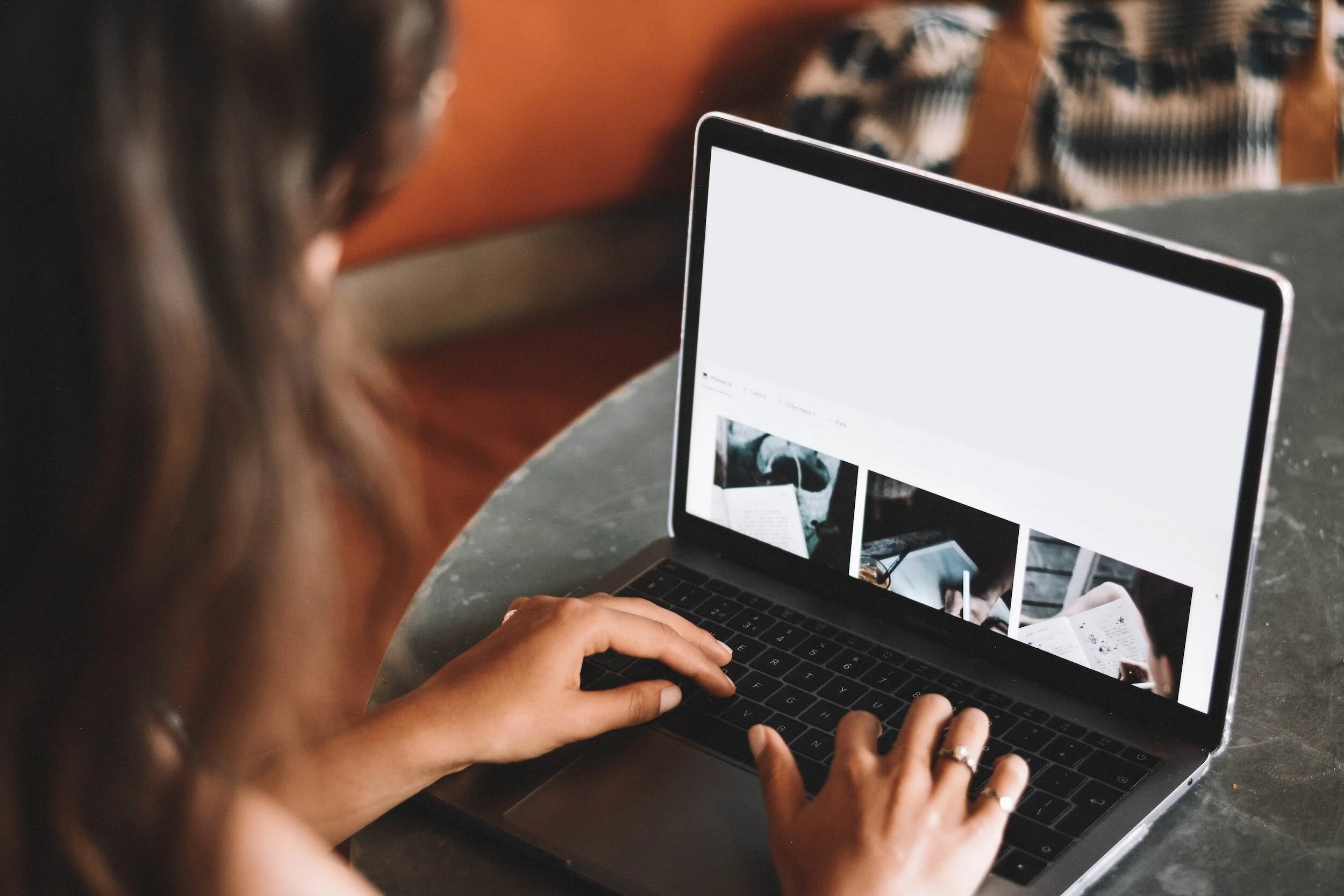 A woman with long brown hair sitting at a table using a laptop to browse a website with photo thumbnails.