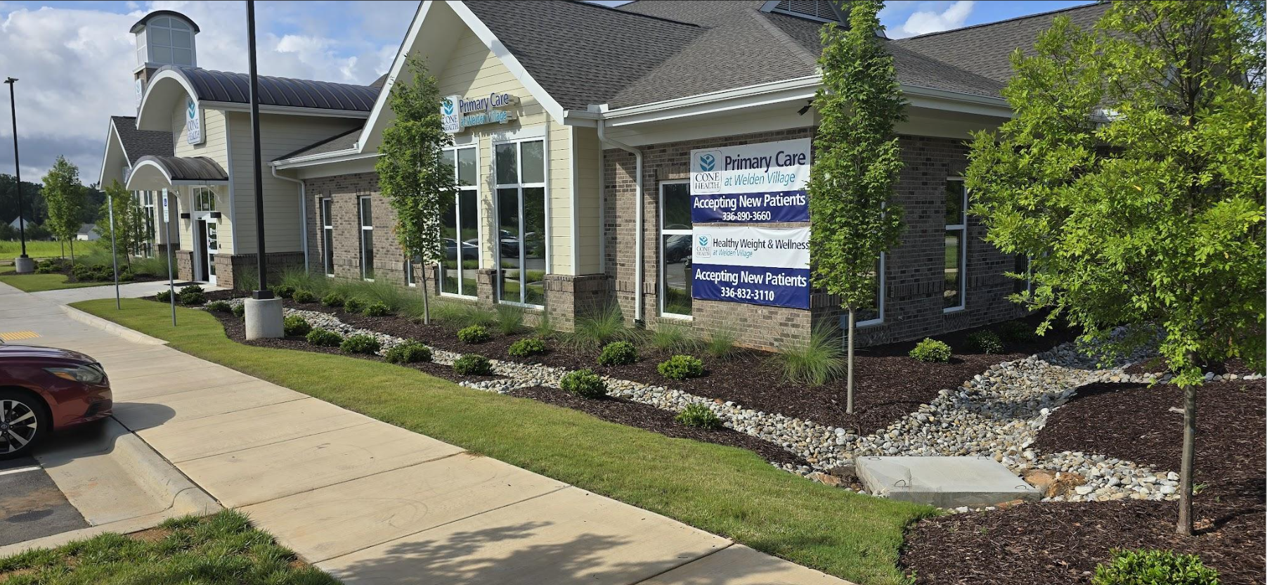 Exterior of a medical clinic building with signs indicating primary care and wellness services, accepting new patients, with landscaped bushes, small trees, and a parking lot in front.