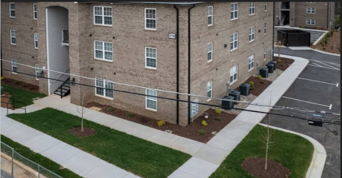 Exterior view of a brick apartment building with sidewalks, small trees, and trash cans outside.