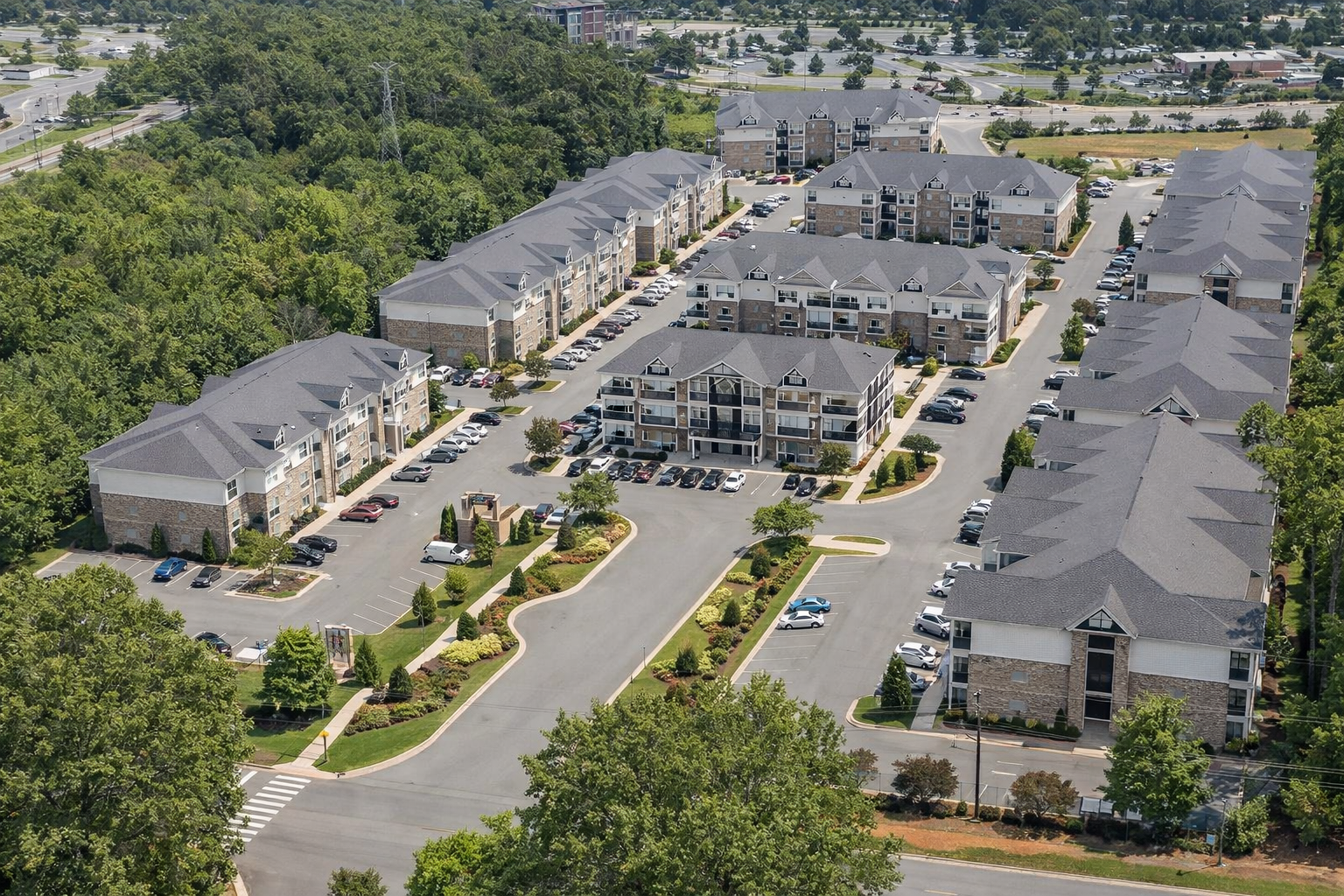 An aerial view of a multi-family apartment complex surrounded by trees and parking lots, featuring beige and gray buildings with dark roofs and landscaped grounds.