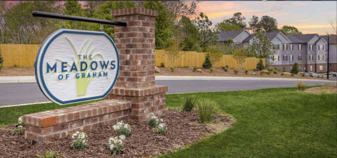 Sign for The Meadows of Graham community with brick pillars and landscaping, residential buildings in the background, during sunset.