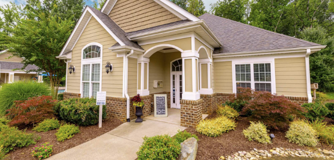 A yellow house with white trim and a brick foundation, featuring a front porch with columns and a small garden with shrubs and flowers.