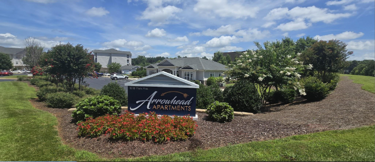 Sign for Arrowhead Apartments in a landscaped area with bushes and trees, apartment buildings in the background under a partly cloudy sky.