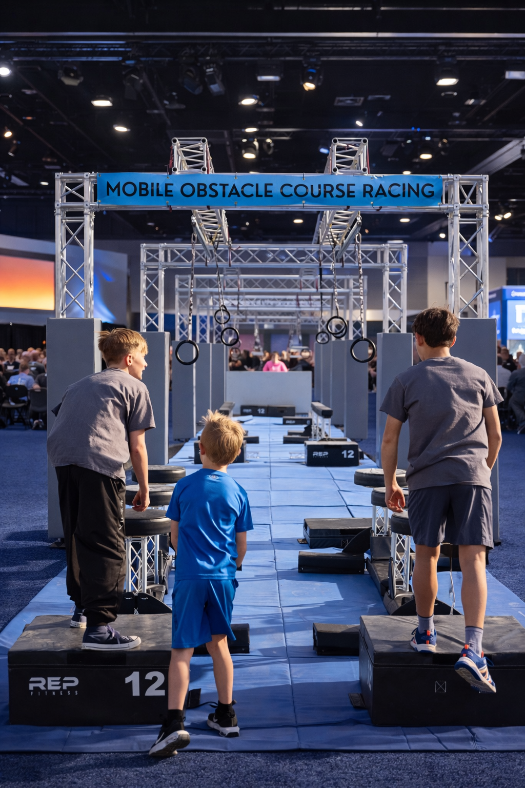 Three boys preparing for a mobile obstacle course race at an indoor event, with rings hanging overhead and a sign that reads 'Mobile Obstacle Course Racing'.