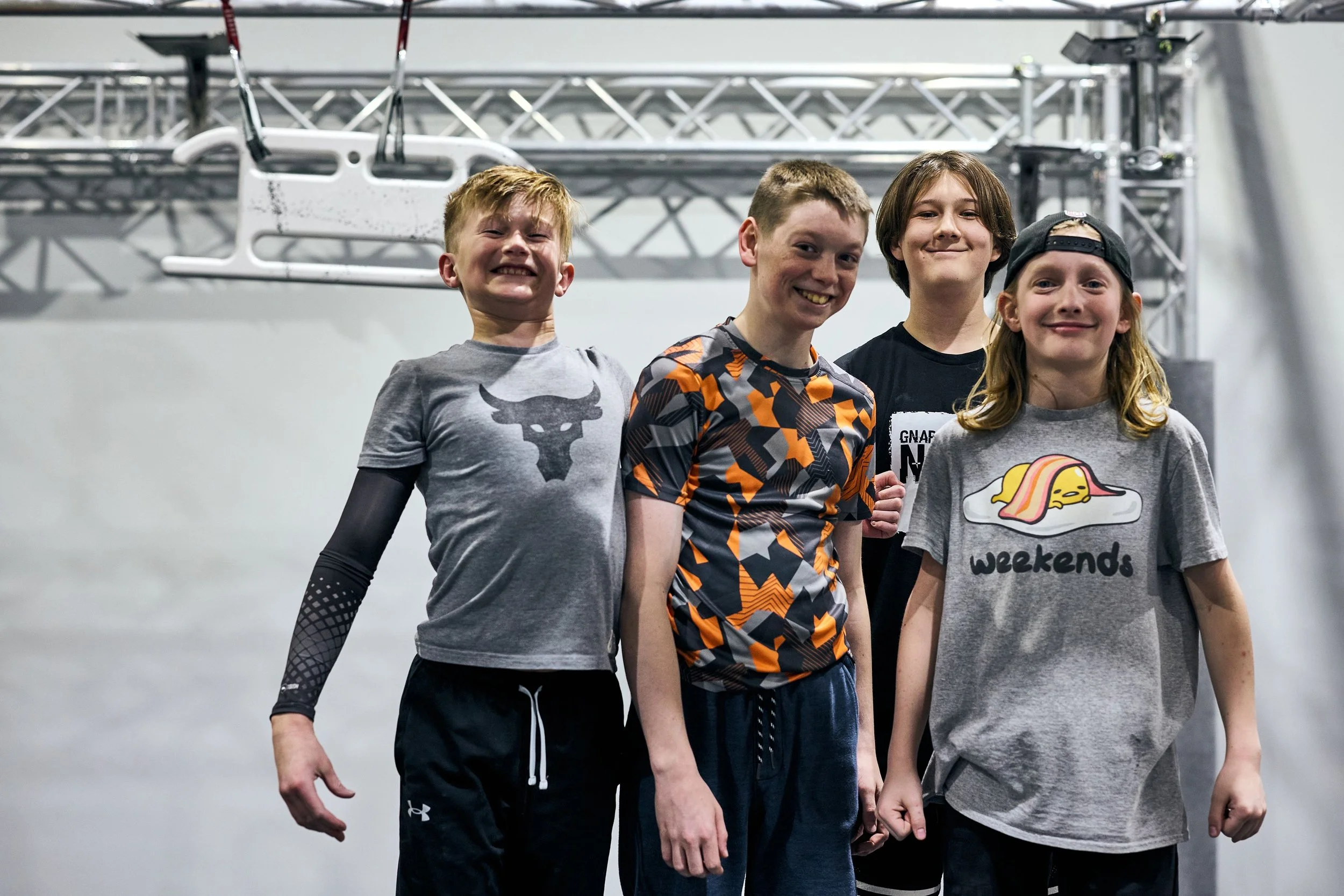 Four boys standing indoors in front of metal scaffolding. They are smiling and dressed in casual and sportswear, with one girl wearing a cap and a T-shirt that says "weekends" and features a cartoon of a sleeping egg.