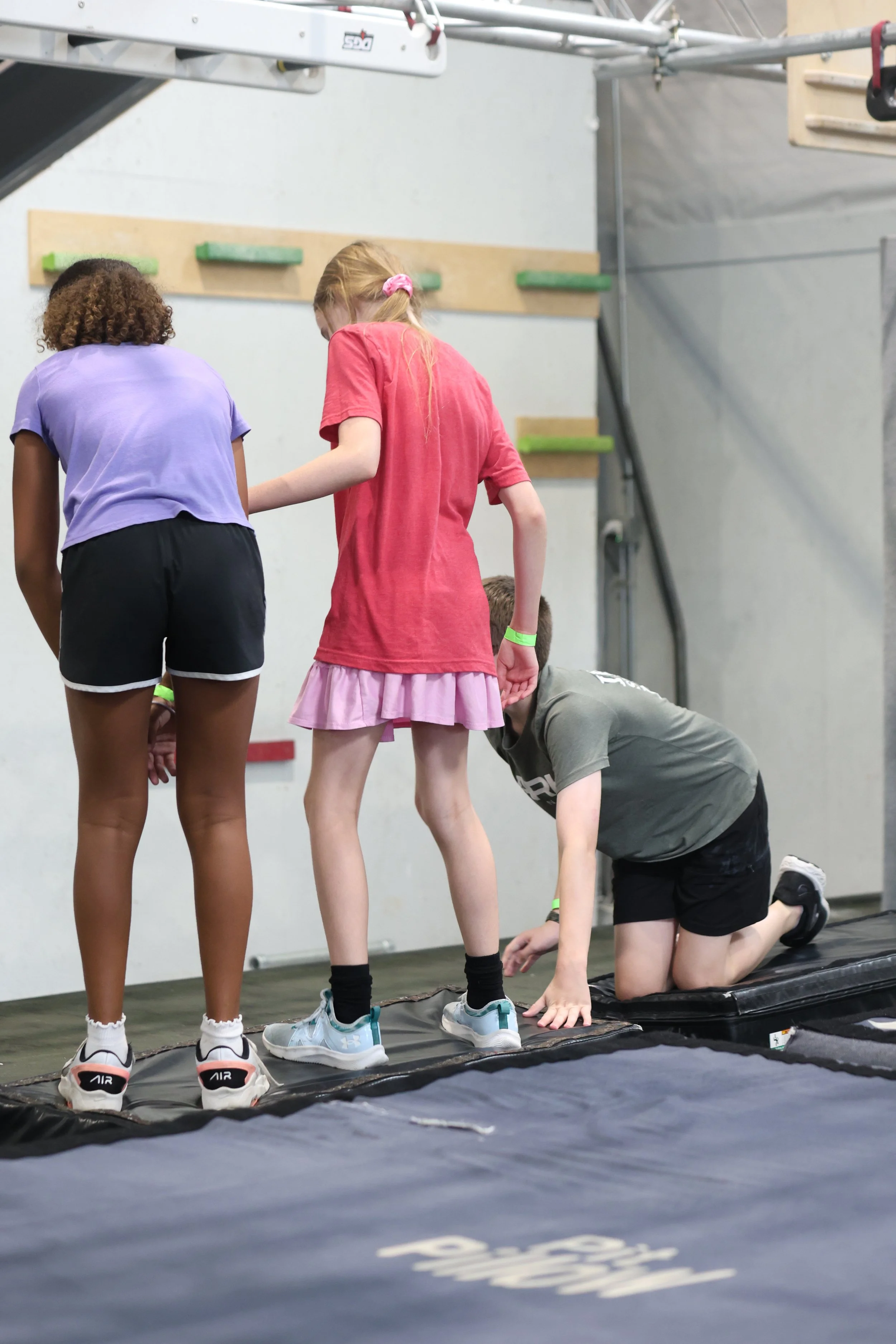 Children practicing parkour on a padded mat in an indoor gym, with some kids observing or assisting.