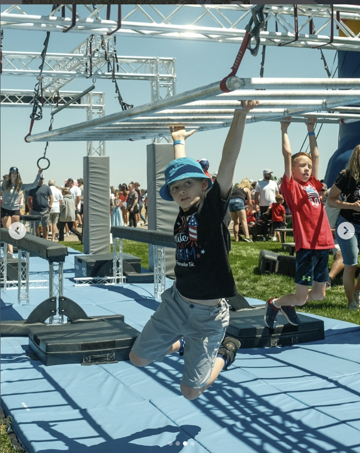 Boy hanging from a metal obstacle during an outdoor event on a bright day, with many people in the background.