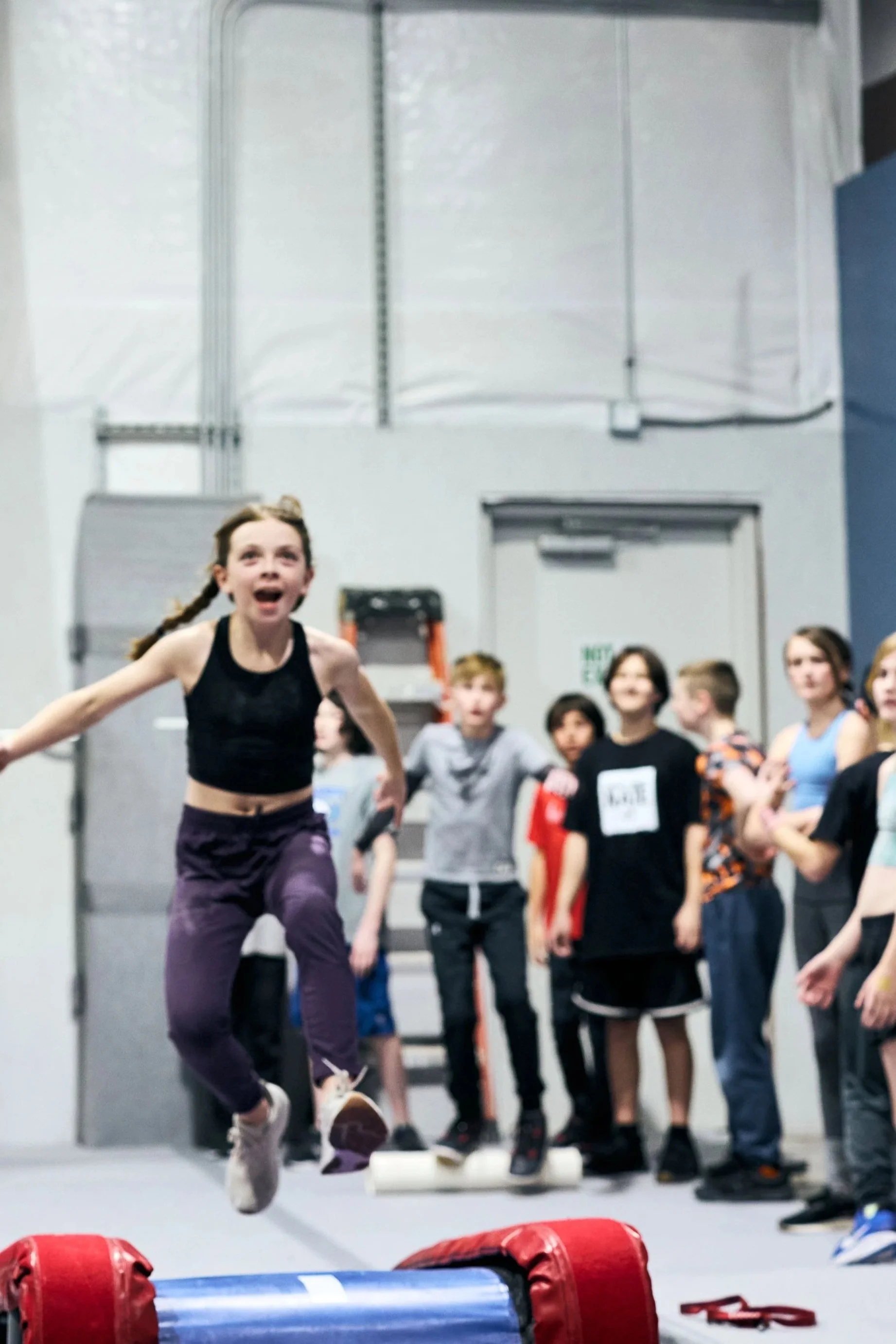 A girl running towards a group of children watching her in a gym or martial arts studio, with punching bags in the foreground and children standing in the background.