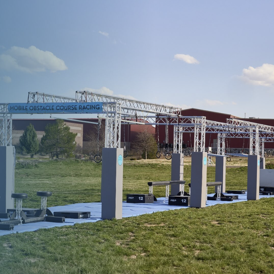 An outdoor mobile obstacle course with metal structures, hanging tires, and gym equipment on a grassy field, under a partly cloudy sky.