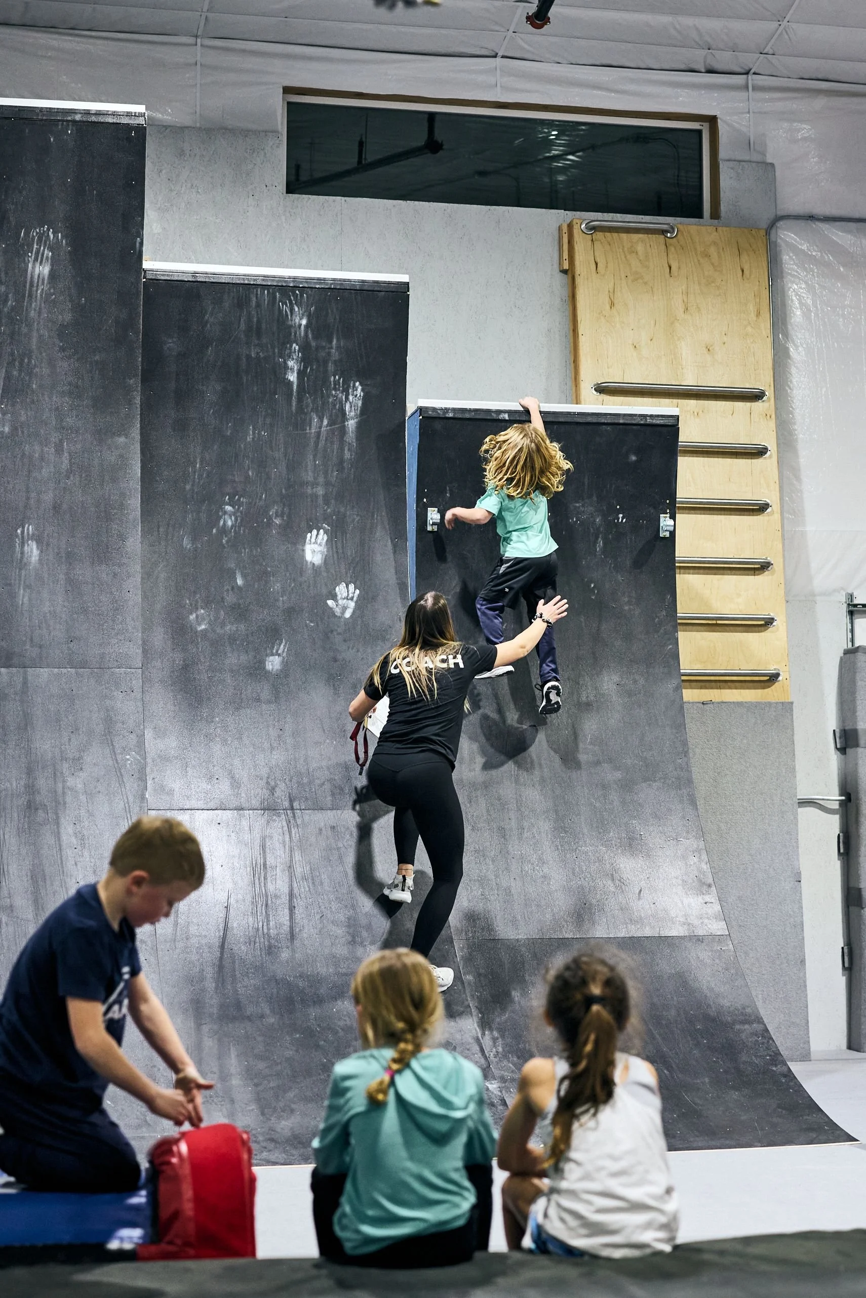 A girl with blonde curly hair climbing a black indoor rock wall with handholds, supervised by a woman, in a gym with multiple children and climbing equipment.