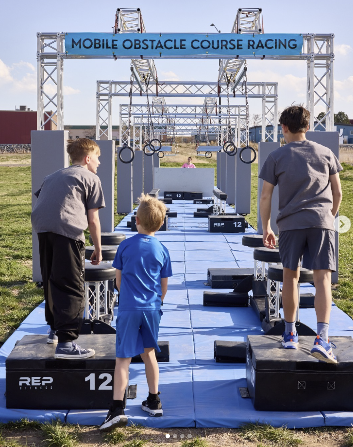 Children participating in a mobile obstacle course race, running across a blue padded pathway with obstacles, rings, and platforms outdoors under a blue sky.