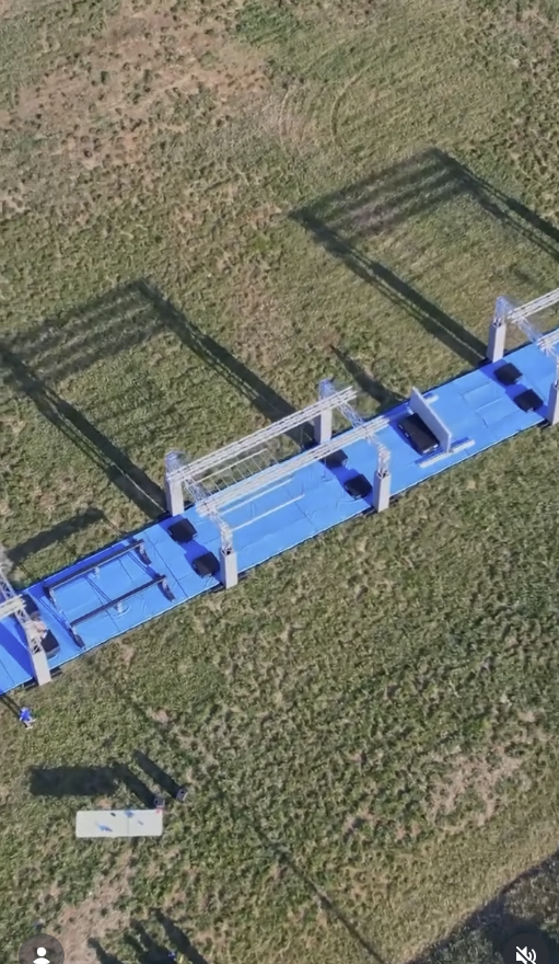 An aerial view of a blue athletic field with empty soccer goals and shadows cast on the grass.