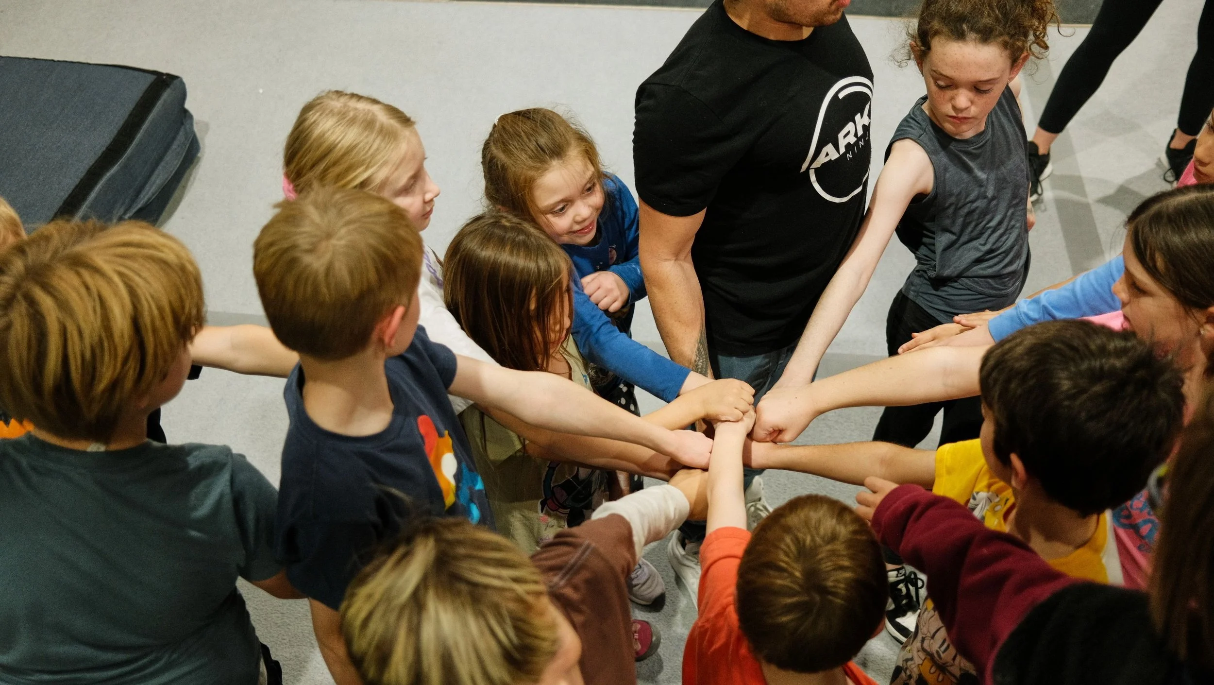 Children and adults in a circle with hands stacked in the middle, participating in a team activity.