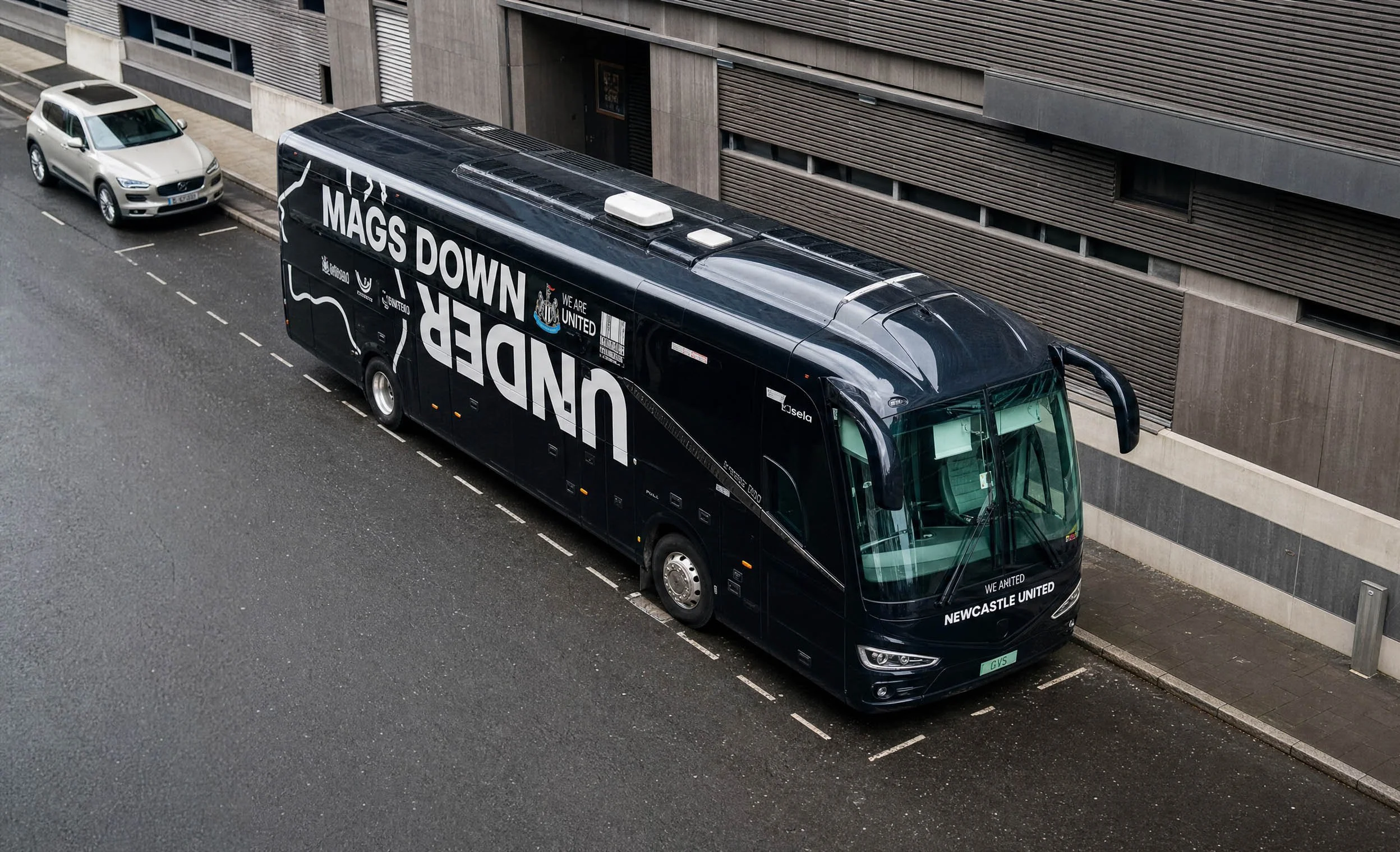A black bus with white lettering parked on a city street next to a gray building. The bus has text reading 'MAGS DOWN UNDER' and 'WE ARE UNITED' with a Newcastle United logo.