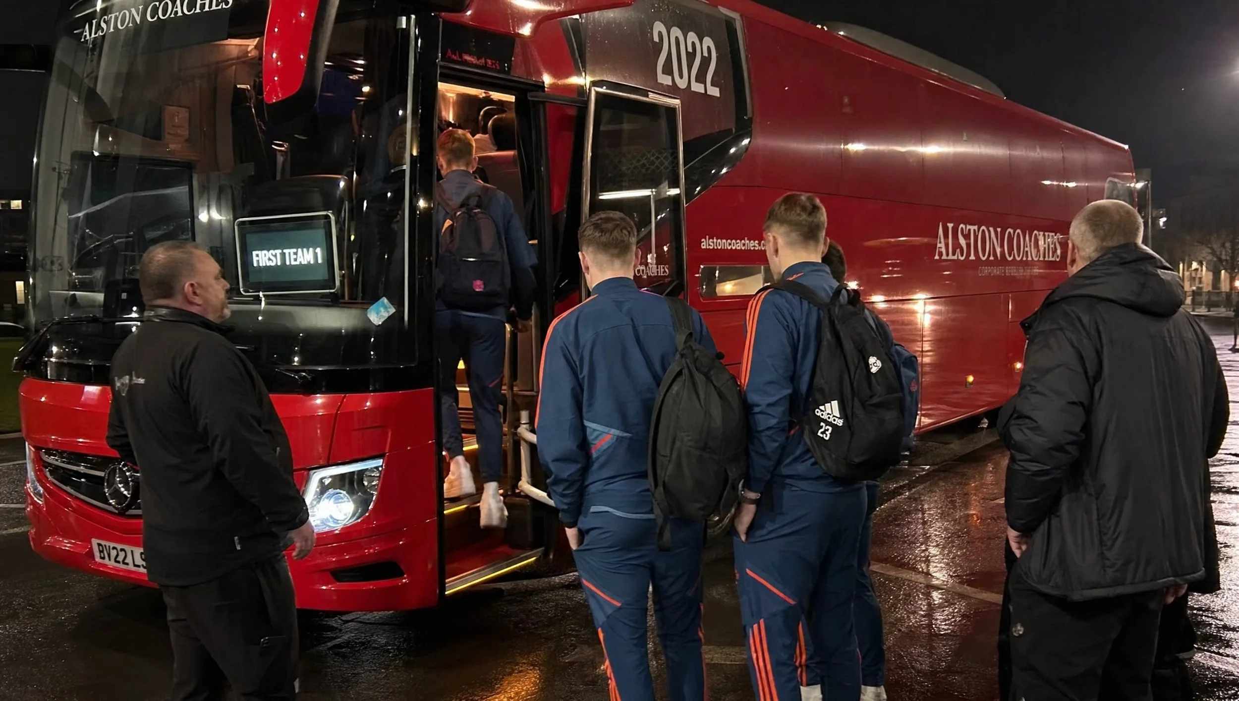 People boarding a red team bus labeled 'Alston Coaches' at night with a sign that reads 'First Team 1'. The bus is outside, and the scene looks wet, suggesting recent rain.