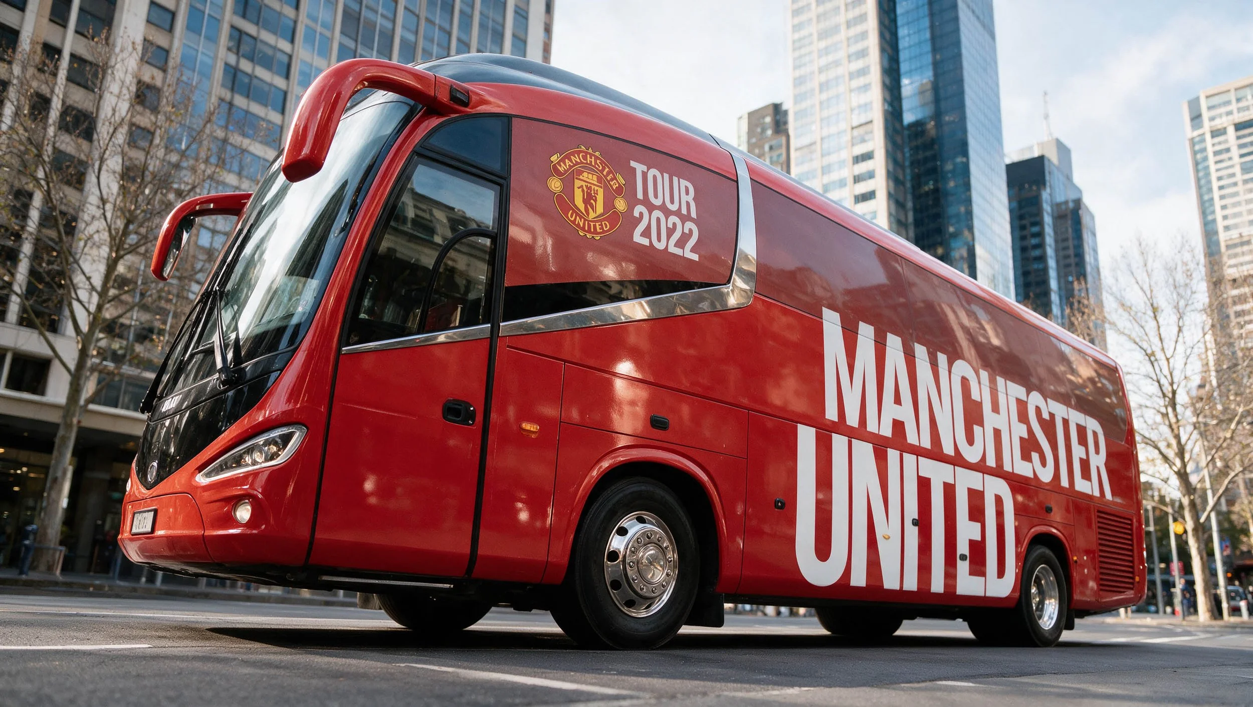 Red Manchester United tour bus parked on a city street with tall glass buildings and leafless trees in the background.