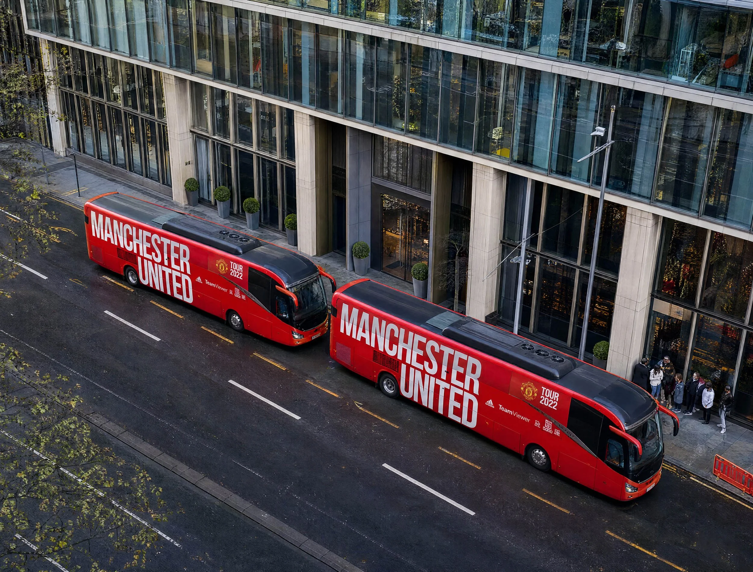 Red tour bus with Manchester United branding parked on city street next to a modern glass building, with people walking nearby and trees lining the sidewalk.