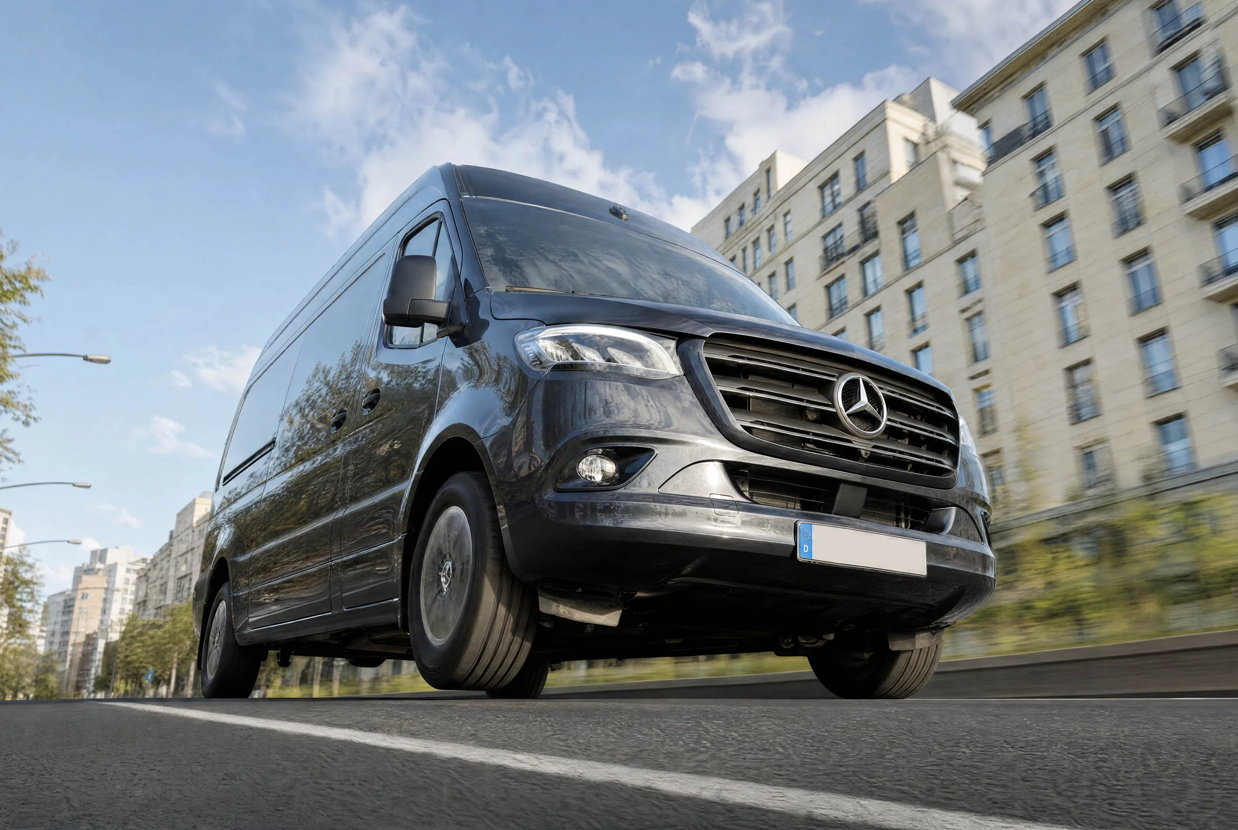 A black Mercedes-Benz van driving on a city street with a building and some trees in the background, viewed from a low angle.
