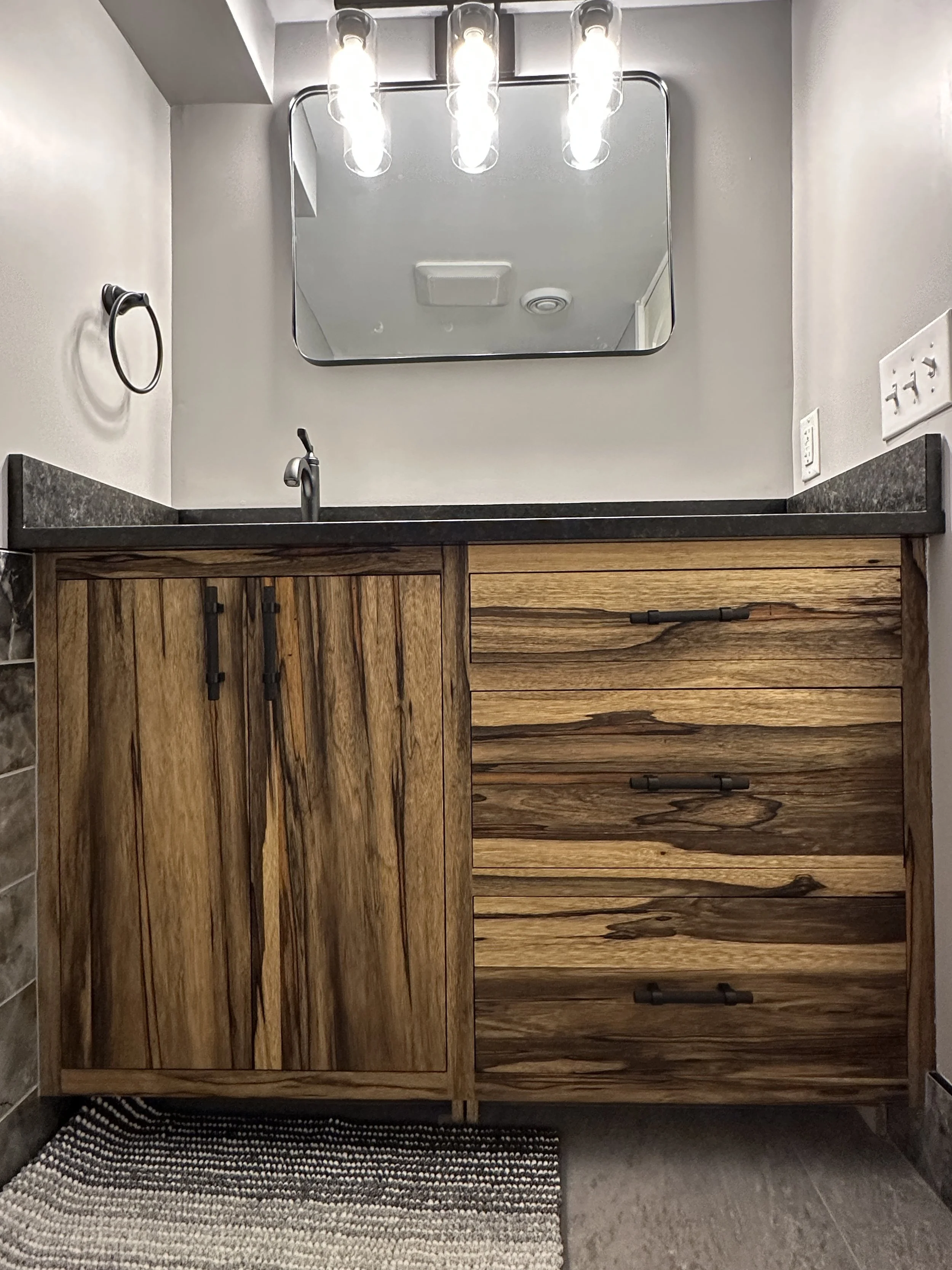 View of a bathroom vanity with a wooden cabinet, black cabinet handles, a black countertop, a sink with a faucet, a rectangular mirror mounted on the wall, a light fixture with three bulbs above the mirror, a towel ring on the wall, electrical outlets, and a textured rug on the floor.