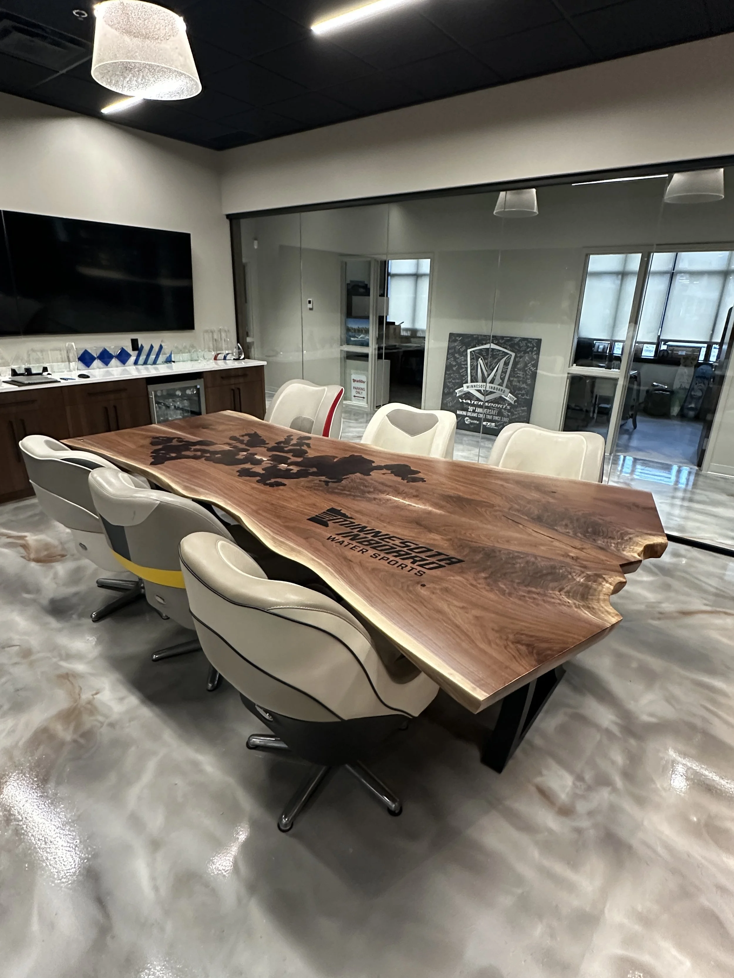 Conference room with a large wooden table, white chairs, a wall-mounted TV, and glass doors leading to an office with posters and windows.
