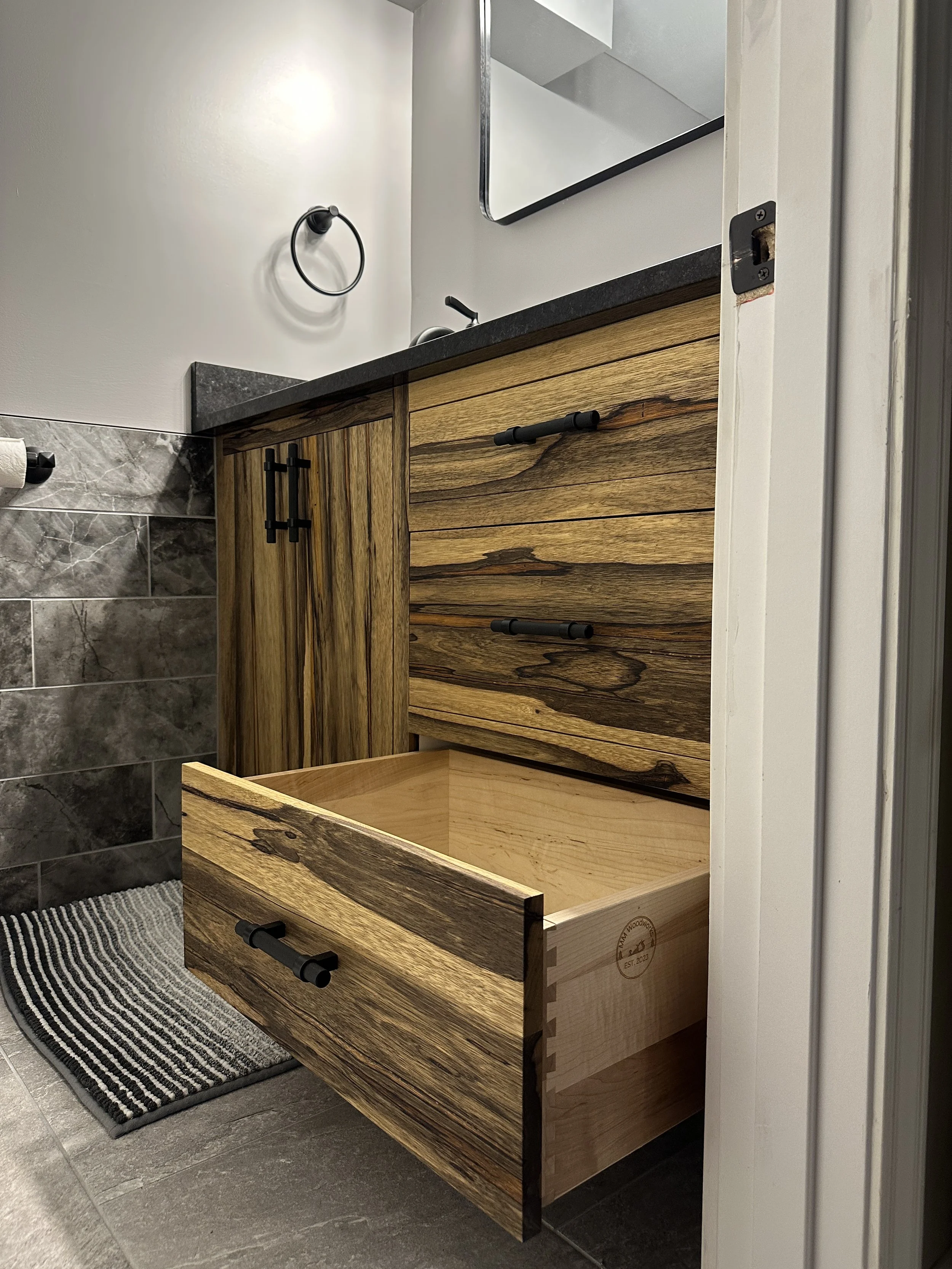 A rustic-style bathroom vanity made of natural wood with black hardware, positioned in front of a white wall and partial gray tiled wall, with a striped black and white rug on a gray tile floor.