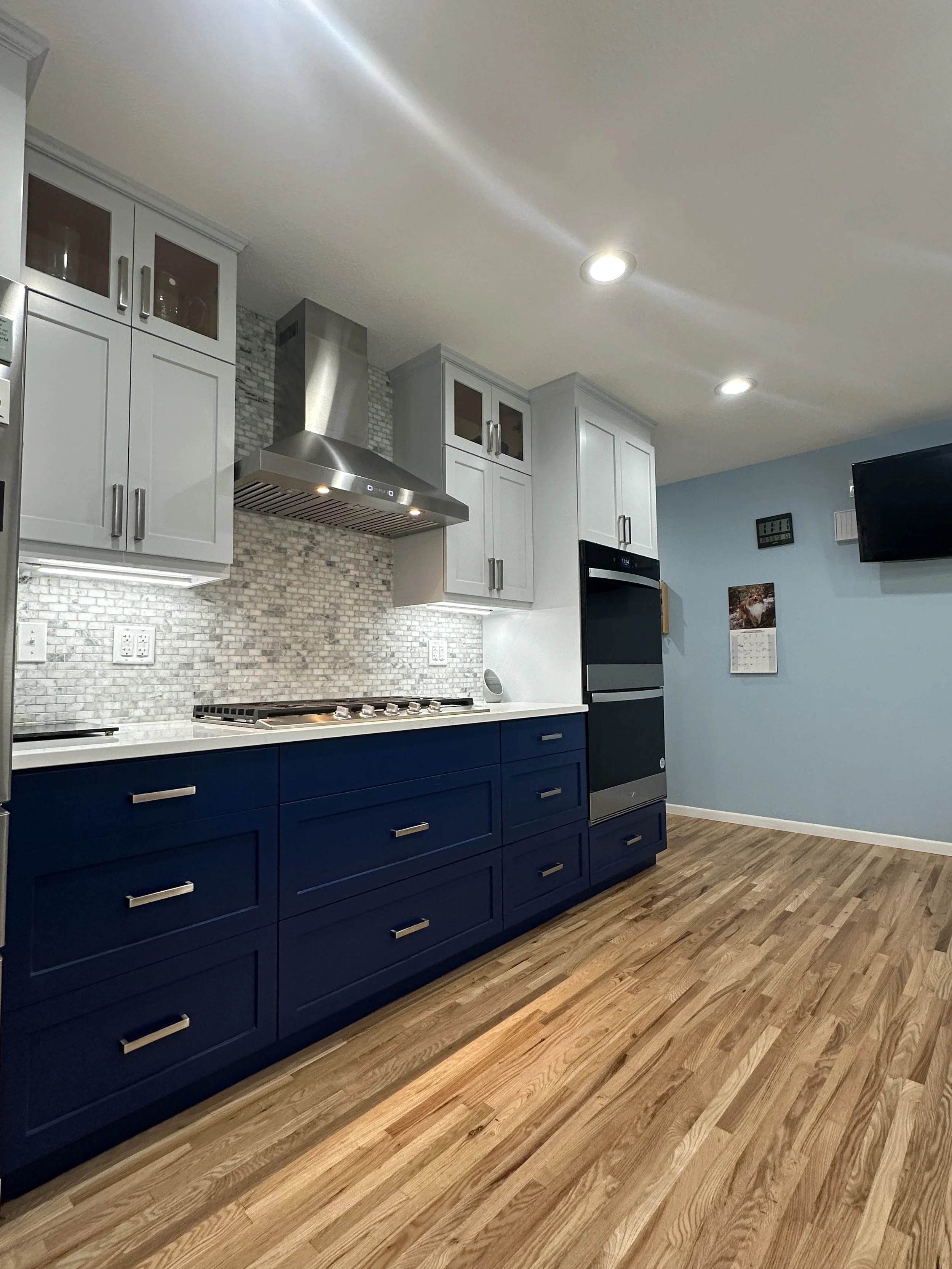Kitchen with white upper cabinets, dark blue lower cabinets, stainless steel stove hood, and wooden flooring.