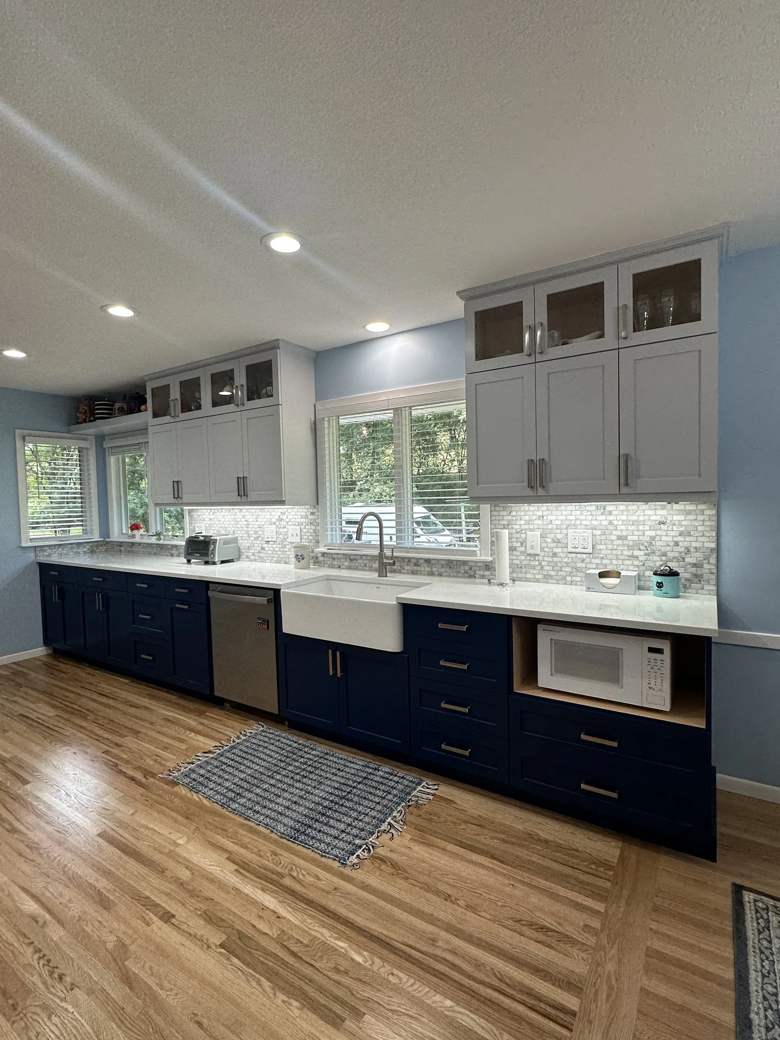 Modern kitchen with blue lower cabinets, white upper cabinets with glass doors, marble countertop, farmhouse sink, and hardwood flooring. Windows with blinds, small appliances, and a woven rug.