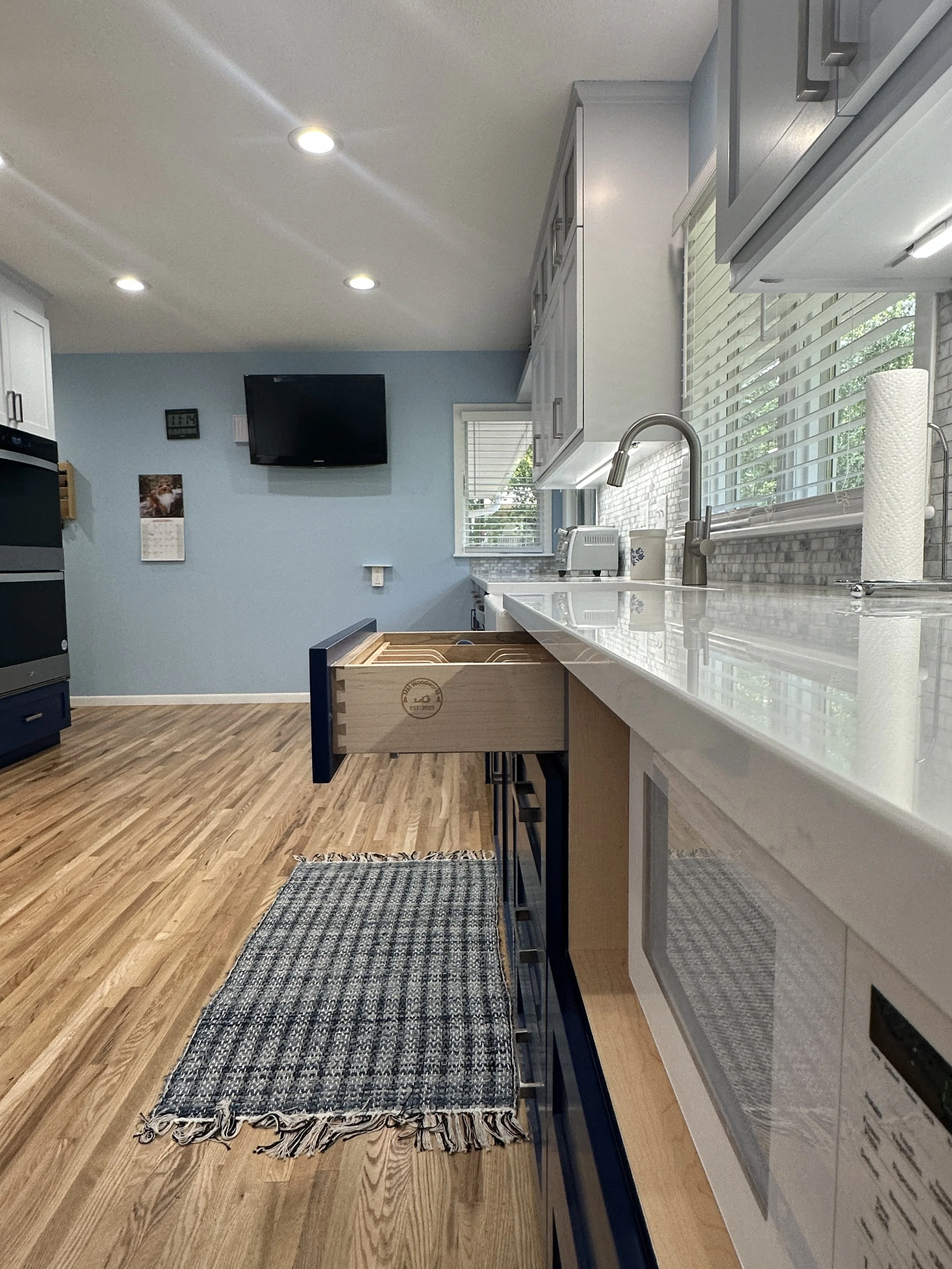 A modern kitchen with white cabinets, a marble countertop, a window with blinds, a view of the outside, a sink with a faucet, a roll of paper towels, a toaster, a cutting board, a blue and beige striped rug, and a wooden drawer partially pulled out. There is a black TV mounted on a blue wall, a small calendar, and a clock.