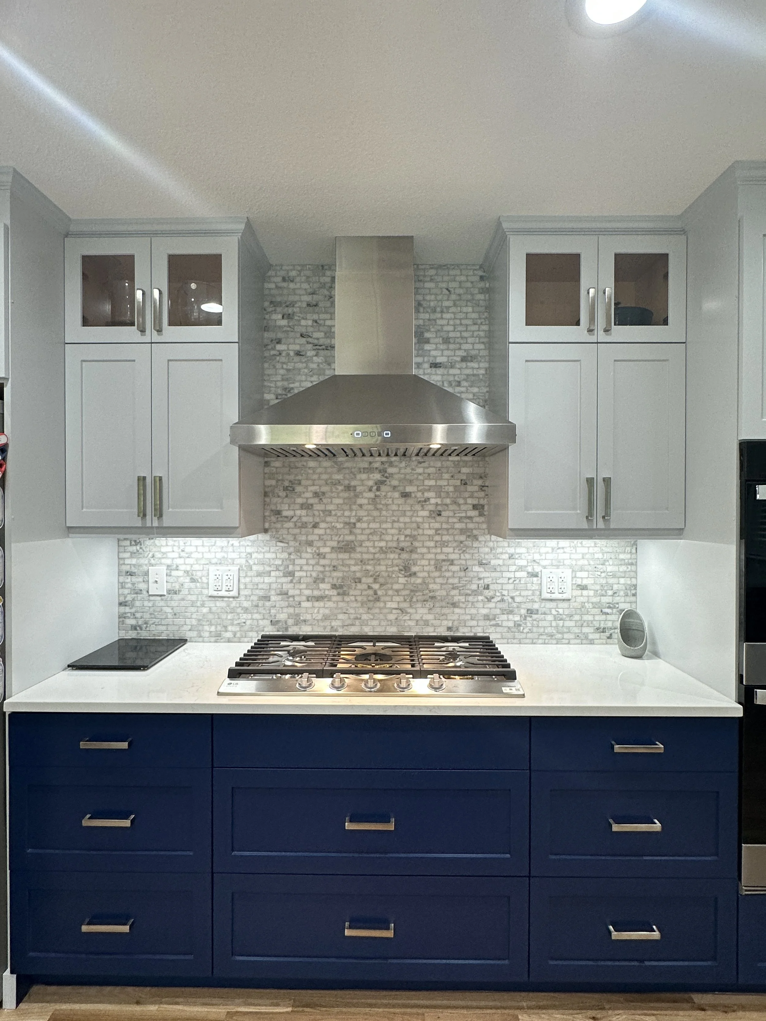 Modern kitchen with white upper cabinets, gray brick backsplash, stainless steel range hood, and navy-blue lower cabinets with silver handles.