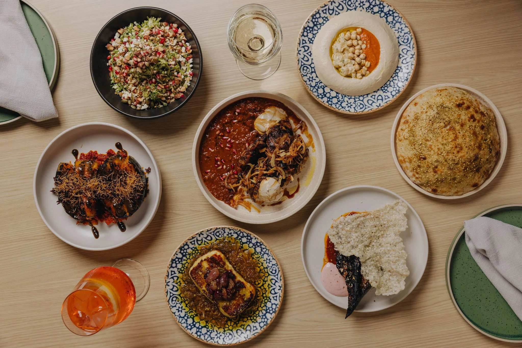 A variety of Middle Eastern dishes on a wooden table, including hummus, a salad with chopped vegetables, a baked dish with tomato sauce, a meat stew with vegetables, and a loaf of bread, with glasses of water and orange-colored beverages.