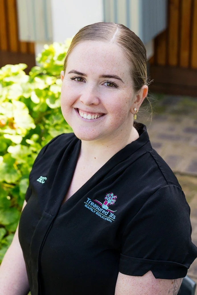 A smiling young woman with blonde hair tied back, sitting outdoors in front of a wooden fence, wearing a navy blue shirt with patches and embroidery.