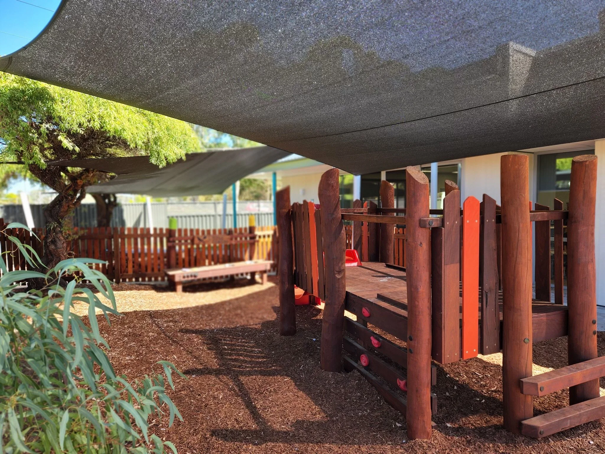 Children's outdoor playground with a wooden play structure, climbing rocks, mulch ground, bench, tree, and shade sail in the background.