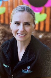 A woman smiling outdoors, wearing a black shirt with a logo that reads 'Treasured Tots Early Education' and a tree graphic, with a background of greenery and trees.