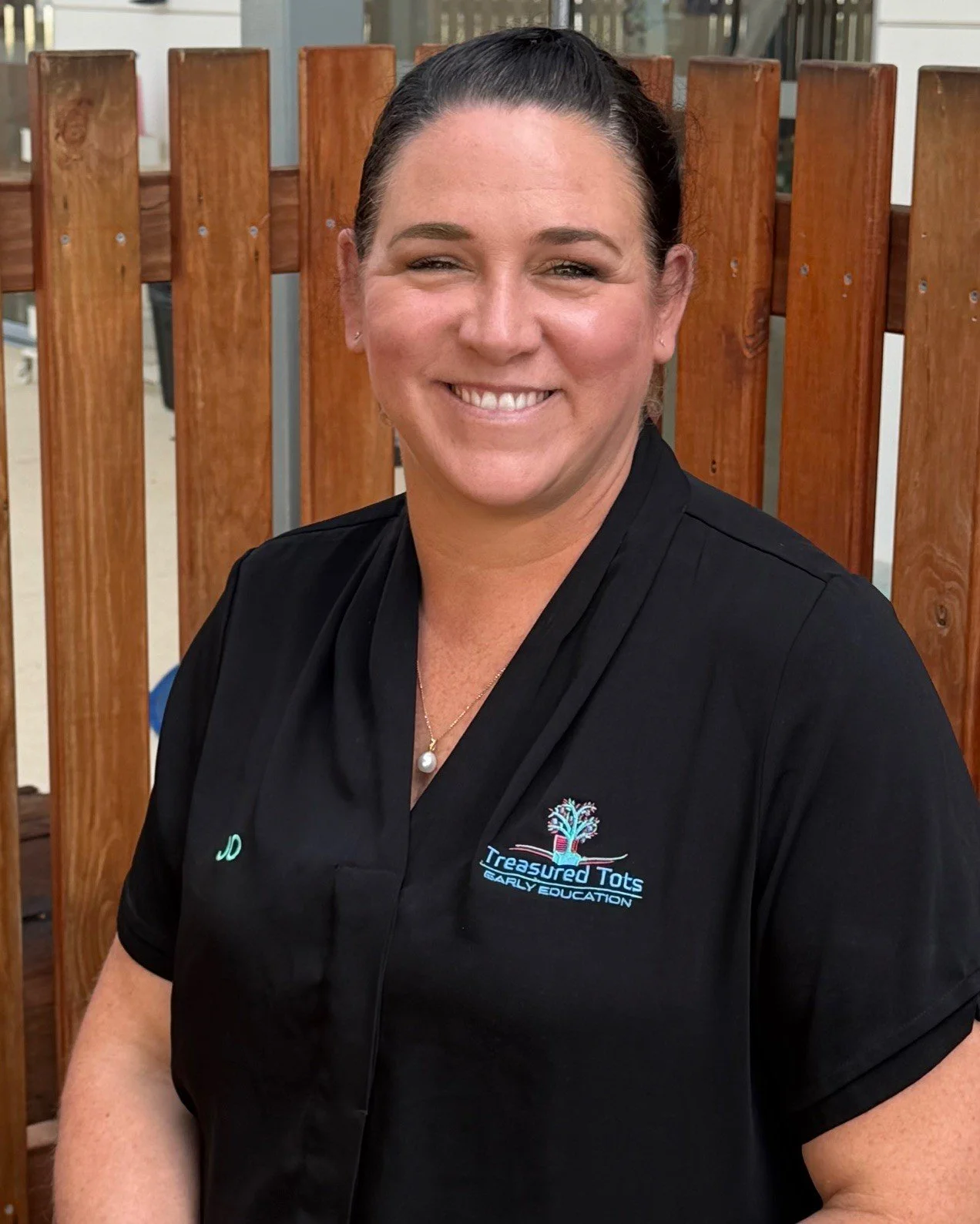 A woman with curly dark hair smiling outdoors next to a wooden fence, wearing a black polo shirt with a logo on the left side.