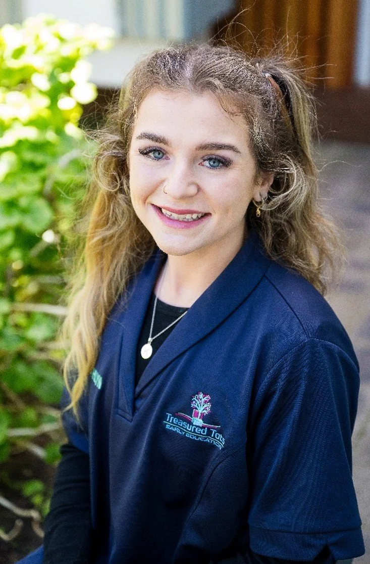 A smiling young woman with blonde hair tied back, sitting outdoors in front of a wooden fence, wearing a navy blue shirt with patches and embroidery.