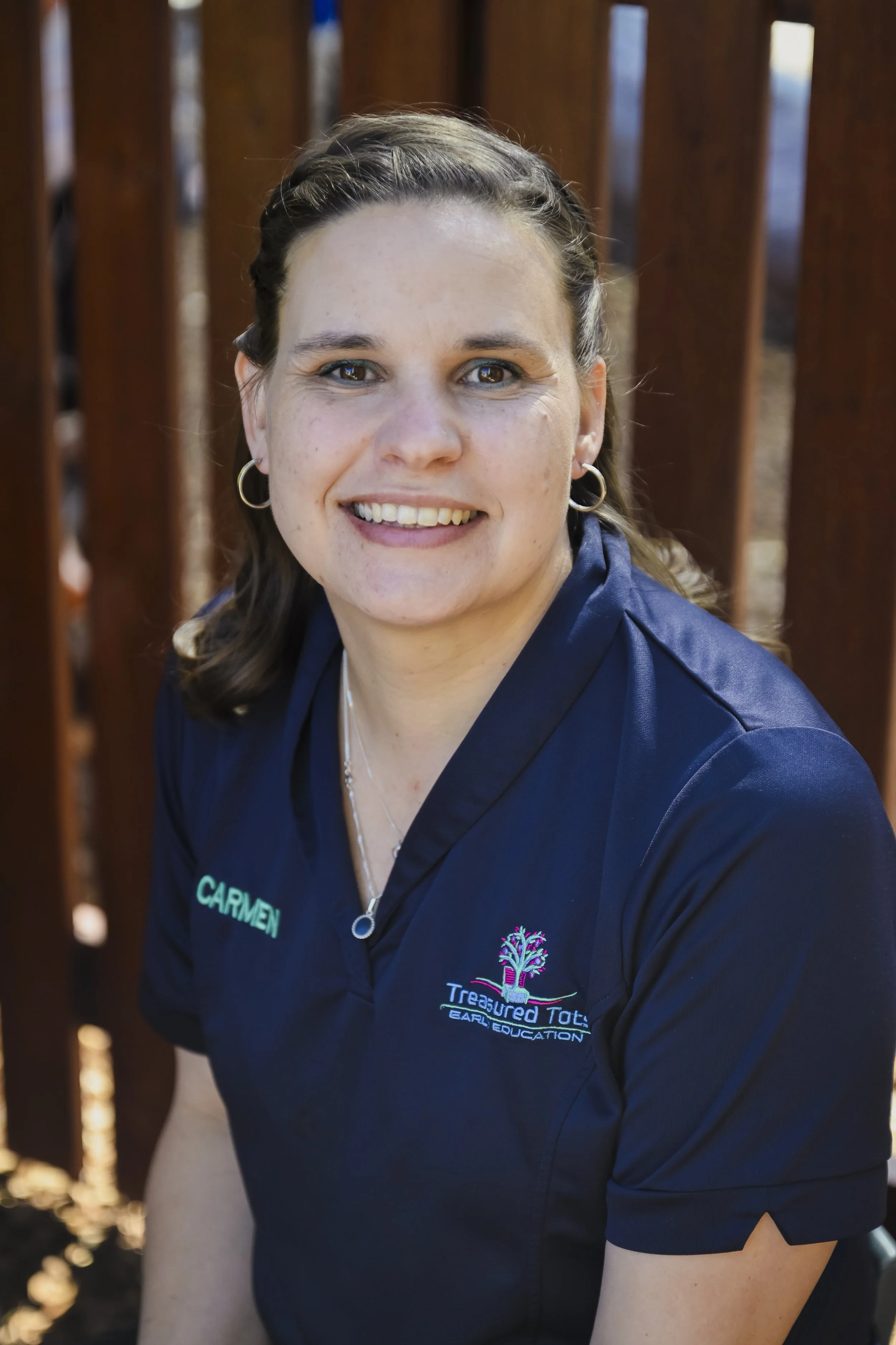 A young woman with light skin and reddish-blonde hair smiling at the camera. She is wearing a navy blue shirt with the words 'SKYE' and 'Treasured Tots Early Education' embroidered on it. She stands outdoors with green foliage and an orange wooden fence in the background.