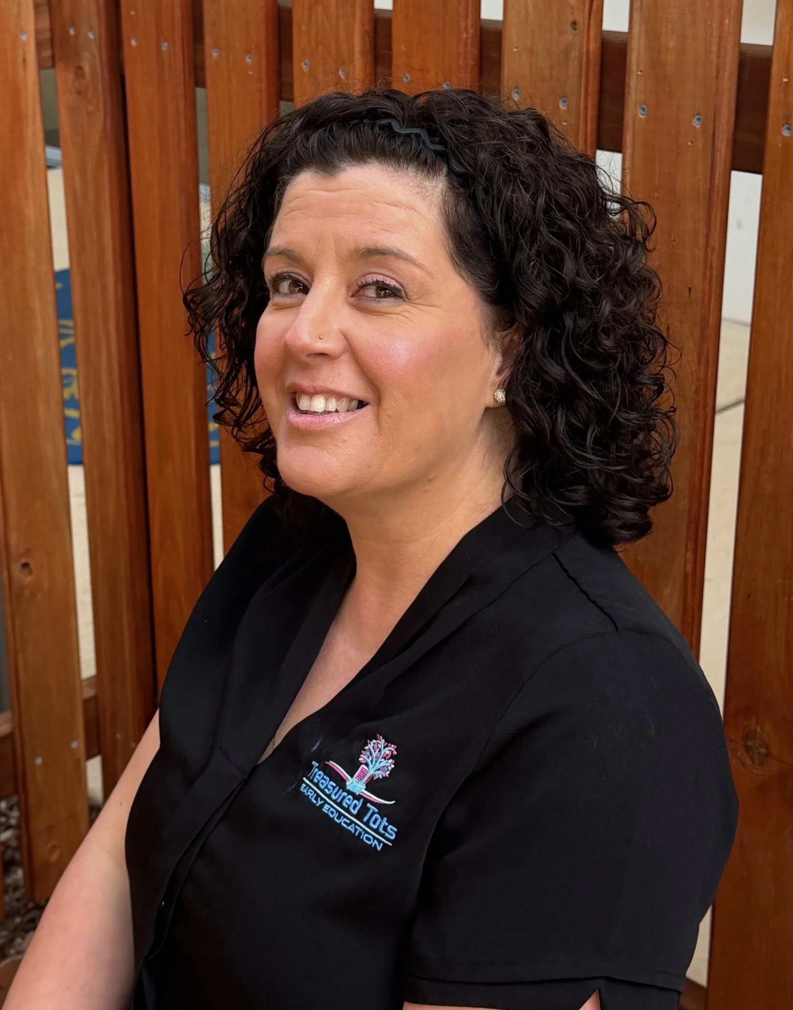 A woman with curly dark hair smiling outdoors next to a wooden fence, wearing a black polo shirt with a logo on the left side.