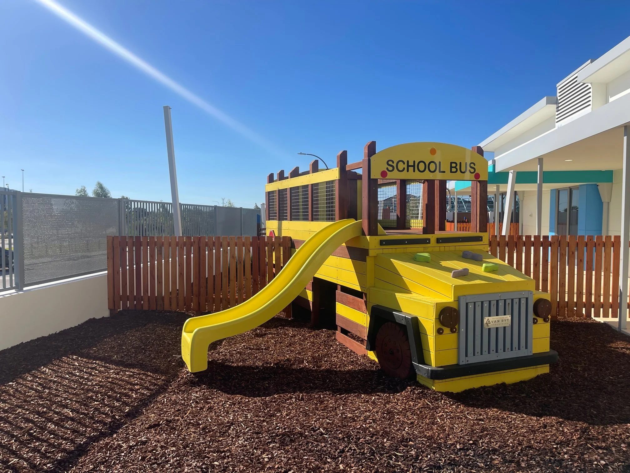 Colorful wooden school bus playground structure with a slide, situated on a mulch surface, surrounded by a wooden fence and modern buildings under a clear blue sky.