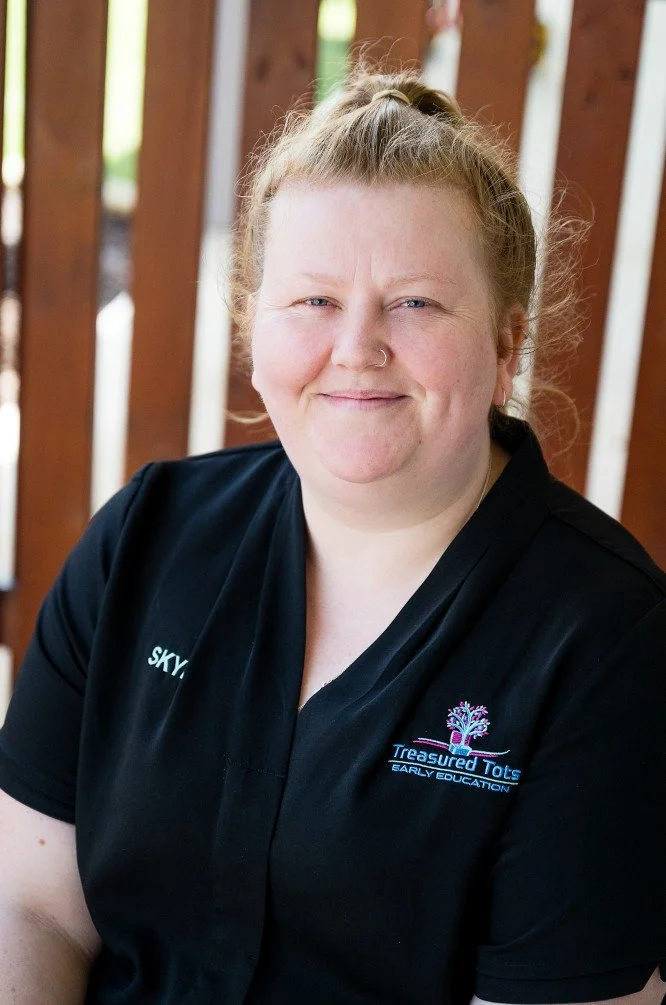 A young woman with light skin and reddish-blonde hair smiling at the camera. She is wearing a navy blue shirt with the words 'SKYE' and 'Treasured Tots Early Education' embroidered on it. She stands outdoors with green foliage and an orange wooden fence in the background.