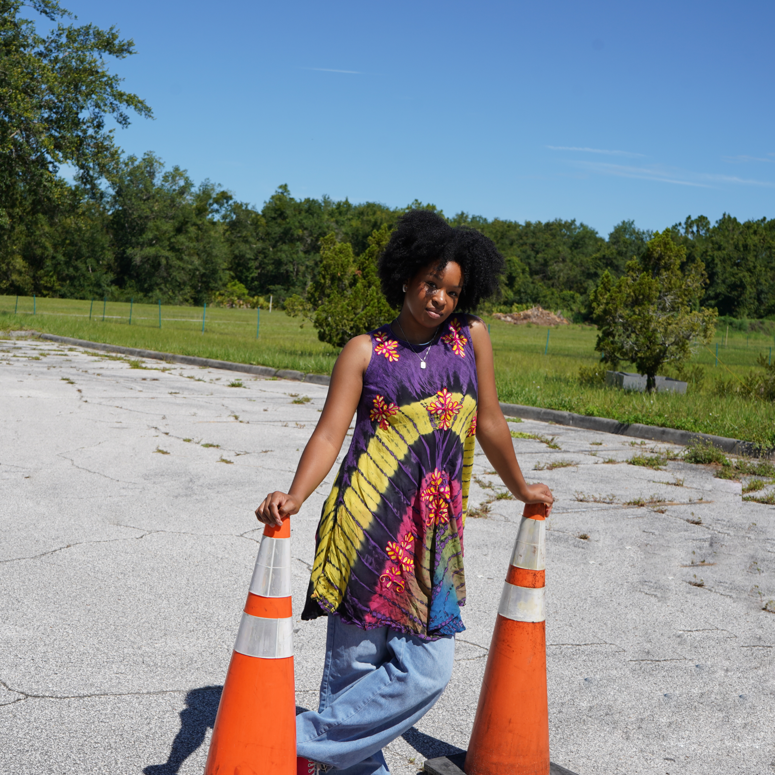 A young woman with curly black hair wearing a colorful tie-dye dress and light blue jeans standing outdoors on a sunny day. She is holding onto orange traffic cones on either side of her, with a grassy area and trees in the background.