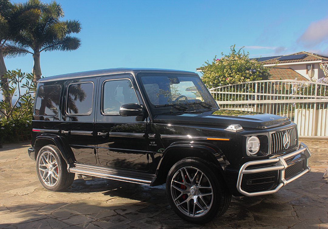 Black Mercedes-Benz G-Class SUV parked in a driveway, surrounded by tropical landscaping and a gated entrance.
